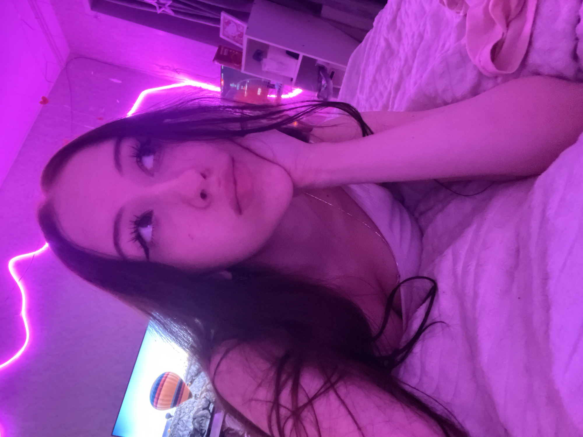 A young woman with long, dark hair is lying on a bed covered with a pink blanket. She is looking up and to the side, with her chin resting on her hand. Her makeup includes dramatic eyeliner and long eyelashes. The room is lit with vibrant pink neon lights, creating a surreal and atmospheric effect. In the background, a television screen shows an image of a hot air balloon. There is also a shelf unit with various items, including books and decorative objects. The overall impression is one of a bedroom setting, possibly decorated with a youthful or trendy aesthetic. The location in Votkinsk, Russia is not discernible from the image itself.