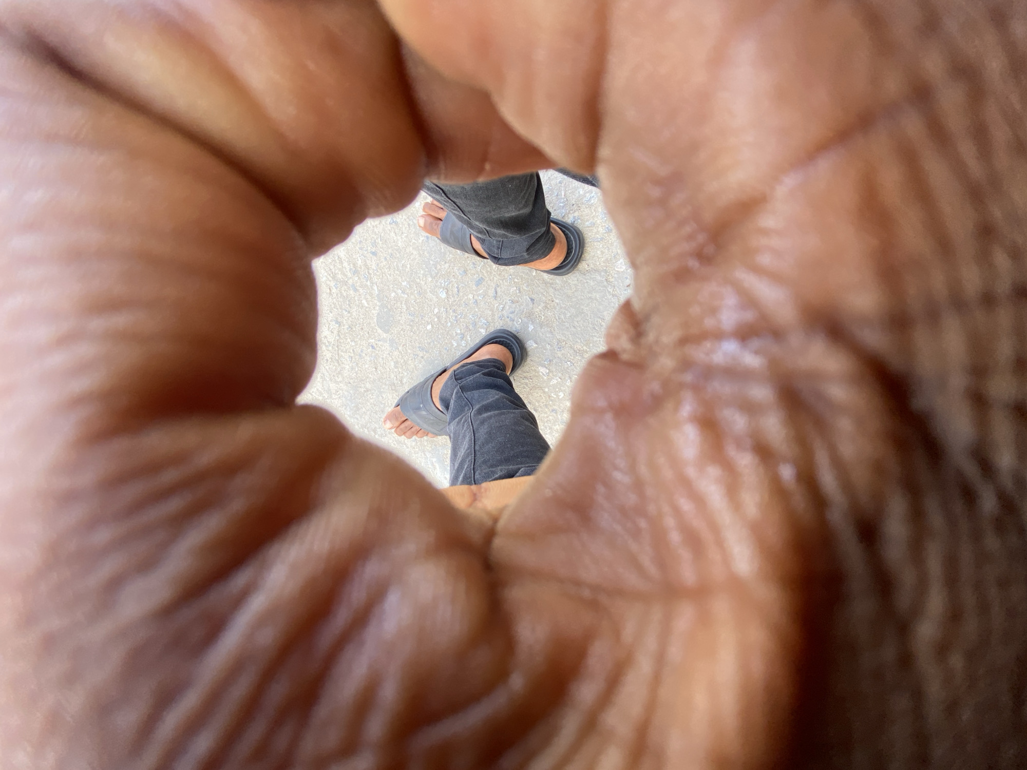 A close-up photograph shows a pair of legs and feet clad in black jeans and black sandals, viewed through a circle formed by human fingers. The ground is light-colored concrete. Location is the city of Ado, Nigeria. There is a person standing on the light-colored concrete and another person looking through their fingers.