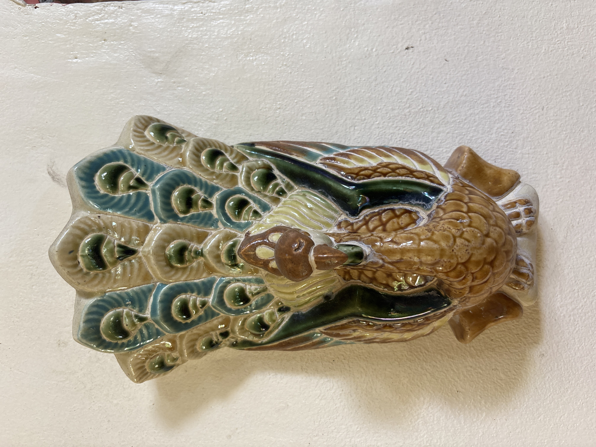 An ornate, glazed ceramic peacock wall plaque is displayed against a textured, off-white wall. The peacock is depicted with its tail feathers fanned out, showcasing a pattern of iridescent blues, greens, and creams. The body of the peacock is a warm brown with detailed feather-like textures. The plaque is positioned on the wall in a well-lit indoor environment, possibly a home or shop, in Chalong, Thailand. There are no people or animals visible in the image. The plaque itself is the sole focus of the photograph.