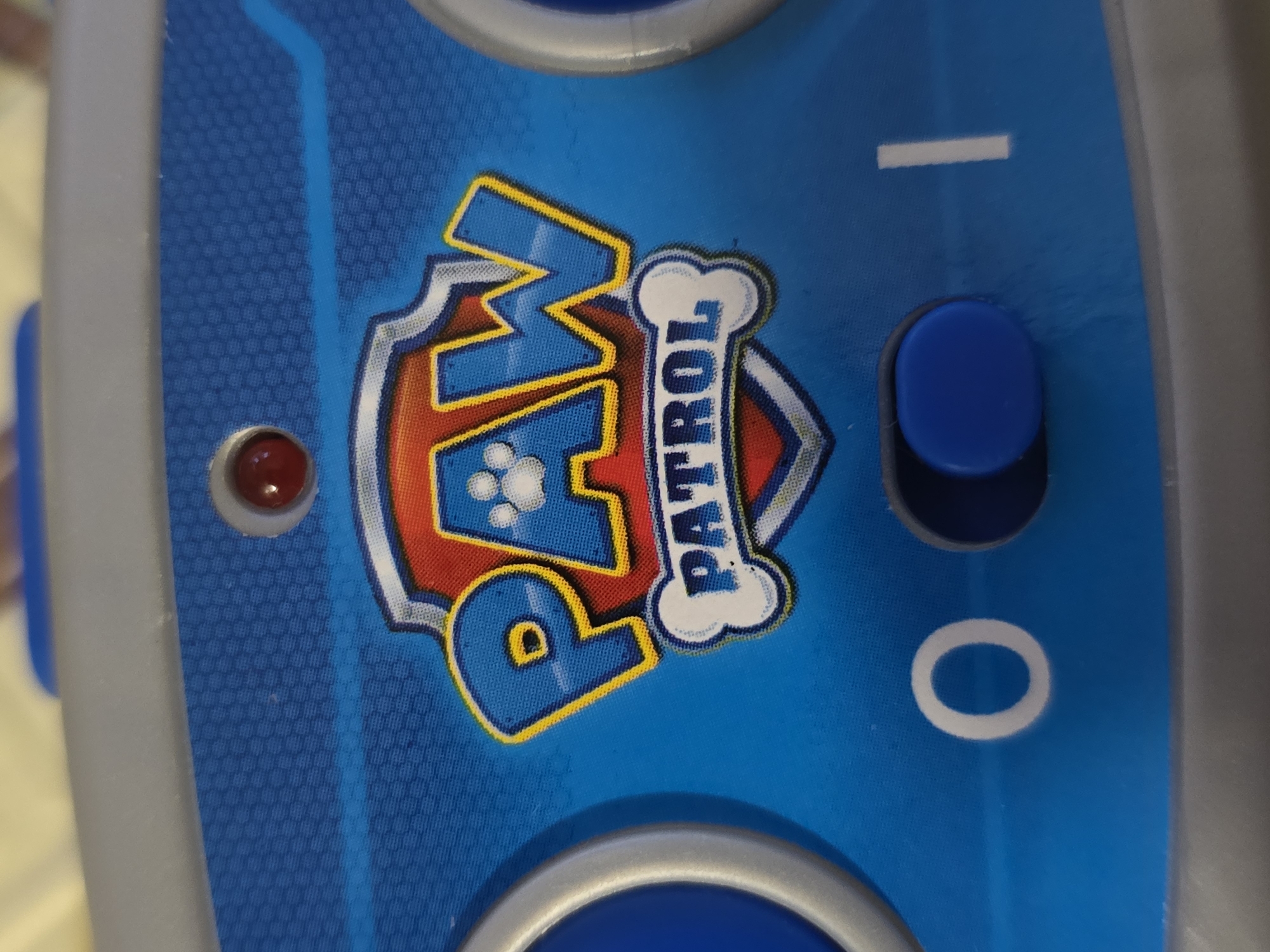 A close-up, slightly angled photograph showcases a section of a blue electronic device, likely a child's toy or controller, framed by a grey/silver plastic casing. The vibrant blue surface is adorned with a subtle, repeating hexagonal texture.
At the center of the image is the prominent "PAW PATROL" logo, rendered in bold red and blue with bright yellow and white outlines. The letter 'A' within "PAW" is creatively replaced by a white paw print, and the word "PATROL" is vertically inscribed on a white bone shape.
To the left of the logo, a small, illuminated red LED light is visible. To the right, a dark blue, oval-shaped button is positioned between two white graphical symbols: a horizontal dash ("-") above it and a circle ("O") below, strongly suggesting an on/off power switch. Another blue circular button, set within a silver ring, is partially visible at the bottom edge of the frame. The background is softly blurred and light-colored, indicating an indoor setting, possibly a tabletop. The image focuses entirely on the device, with no human subjects or specific activities depicted, emphasizing the branding and interface of the toy.