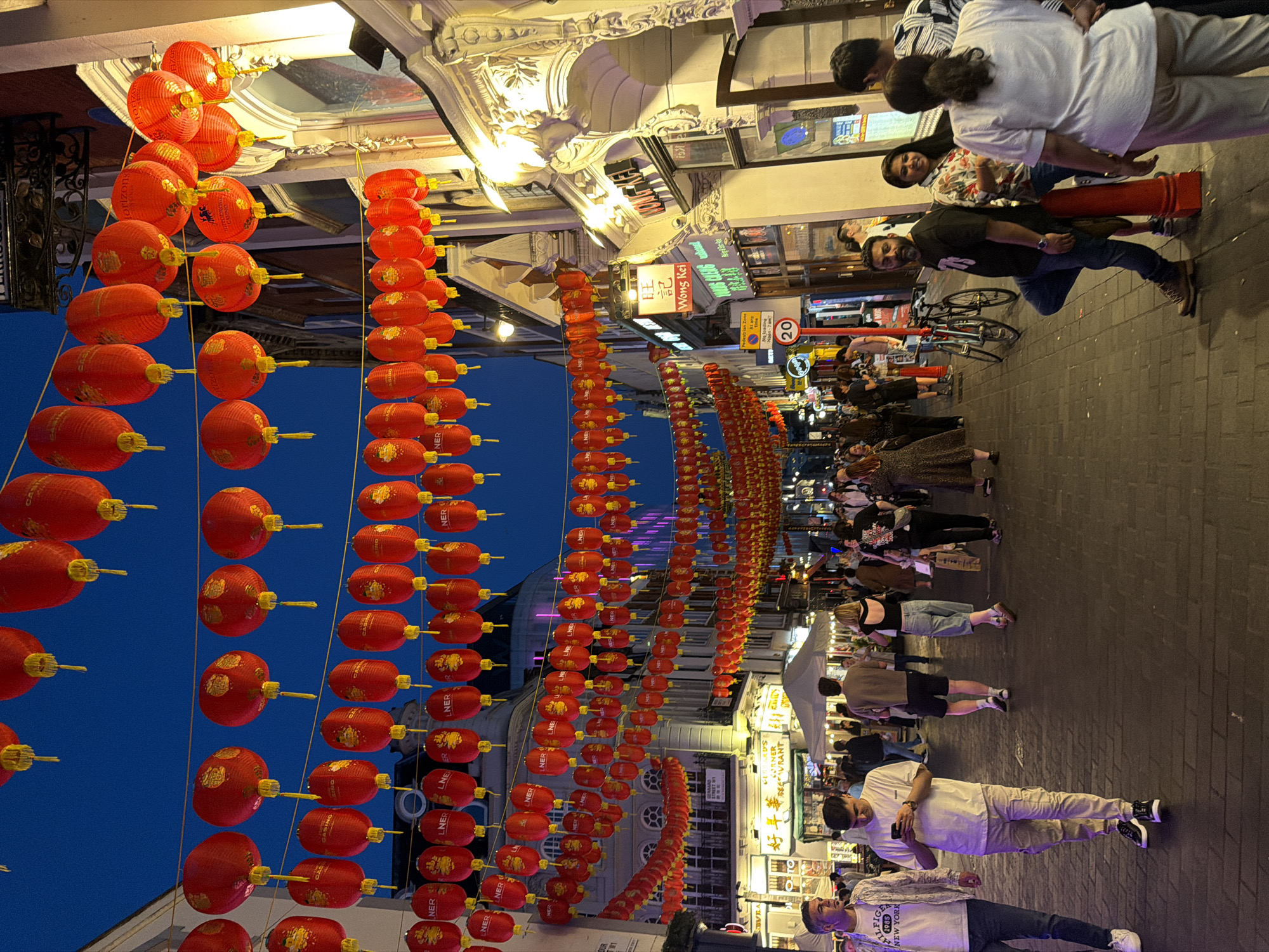 This is a street scene in Chinatown, London, United Kingdom, at dusk. Rows of red paper lanterns line the street, hung overhead. People are walking along the brick pavement. There are shops and restaurants visible along the sides of the street, with signs in English and Chinese. One sign reads "Wong Kei." A few people are standing and talking or looking at their phones. There is a 20 mph speed limit sign visible.