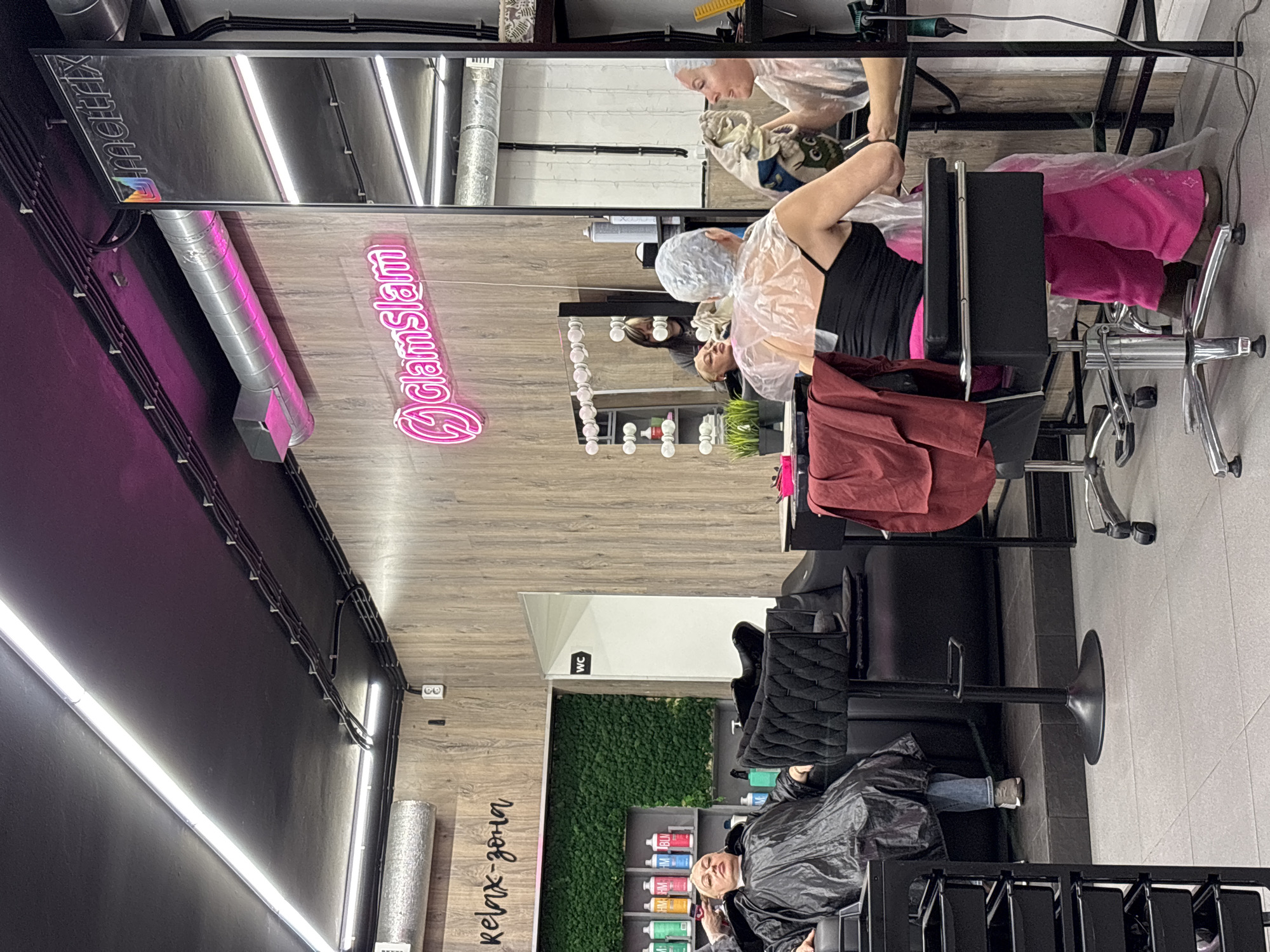 The image shows the interior of a hair salon. Customers are sitting in salon chairs, likely undergoing hair treatments, as evidenced by the hairnets on their heads and protective coverings around their shoulders. One customer is lying down in a reclined chair in what appears to be a "relax-zona" or relaxation zone. The salon features a modern design with neon signage, a vertical garden wall, and metal ventilation ducts. Mirrors reflect the scene, creating the illusion of a larger space. The location is likely in the city of Saint Petersburg, Russia, based on the Cyrillic text visible in the photo.