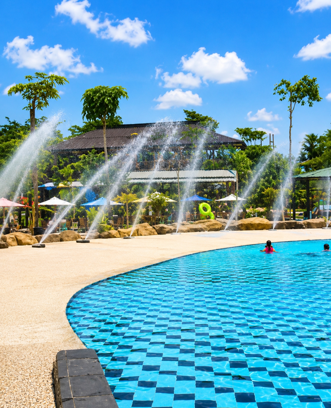 In the city of Eampat Kwalamencirim, Indonesia, a bright, sunny day is depicted at a recreational water park. The foreground is dominated by a large, curving swimming pool with a blue and black checkered tile pattern. The water is clear and reflects the bright blue sky dotted with fluffy white clouds. Two individuals are visible in the pool; one, a child in a pink vest, is swimming, and another person is partially visible further away.
The pool deck is a light tan color with a textured finish. Along the edge of the pool, a darker, brick-like border is present. Beyond the pool deck, a landscaped area features rocks, palm trees, and lush green foliage. A series of tall water fountains are spraying arcs of water into the air, creating a misty effect against the backdrop of trees and a dark-roofed building. Several umbrellas, some white and some blue, are set up in the area, suggesting seating or shaded spots. A bright green inflatable ring is also visible on the ground near the fountains. The overall scene conveys a sense of leisure and summer fun.