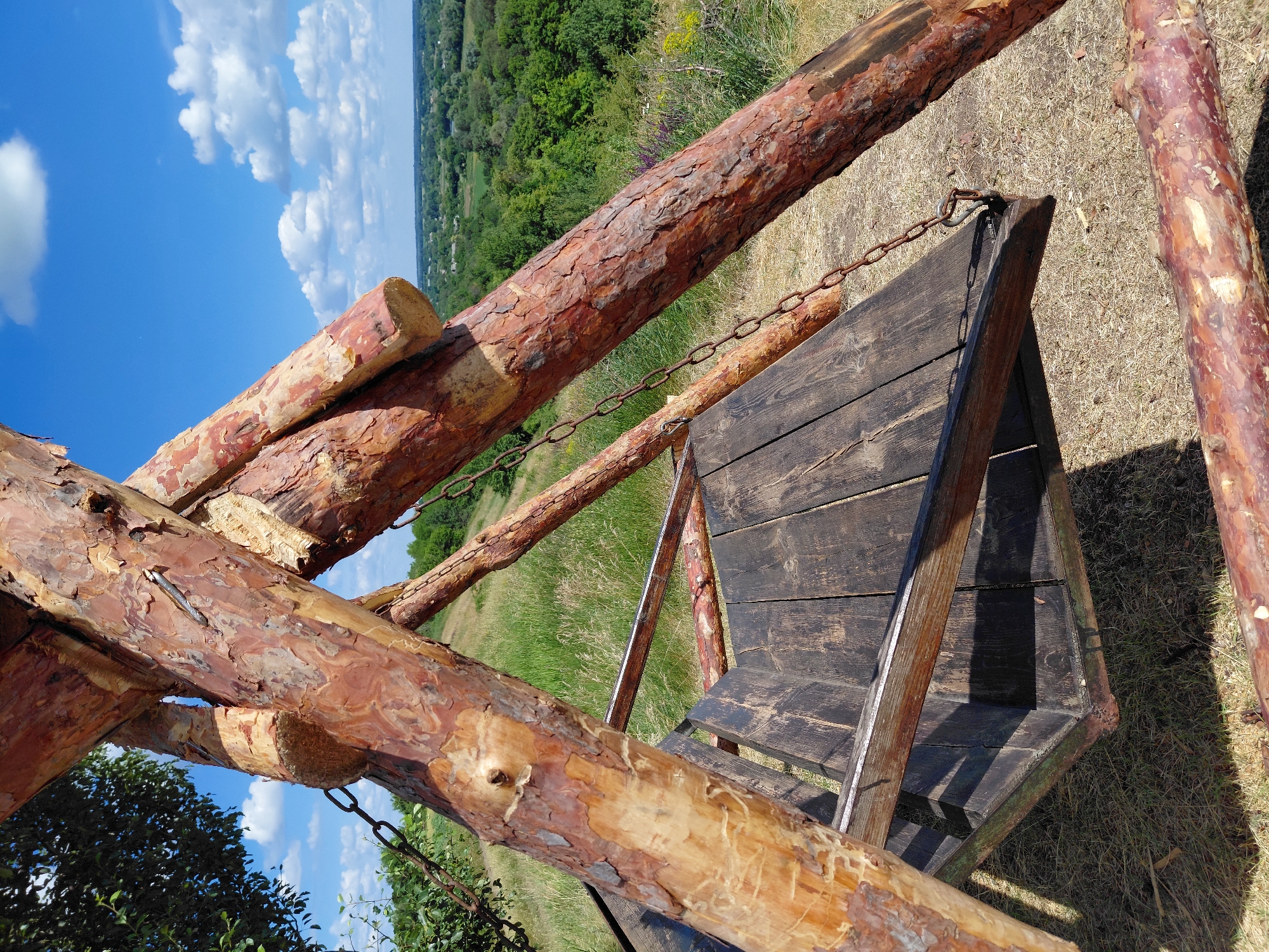 A rustic, wooden swing set made of rough-hewn logs is situated on a grassy hillside under a bright blue sky with scattered white clouds. The swing seat is constructed from dark, weathered wooden planks. The supporting structure is made of large, natural logs with visible bark. Chains connect the seat to the overhead frame. In the background, a lush green landscape of trees and rolling hills stretches towards the horizon. The sun casts shadows on the wooden seat. The setting appears to be rural and outdoors, away from an urban environment.