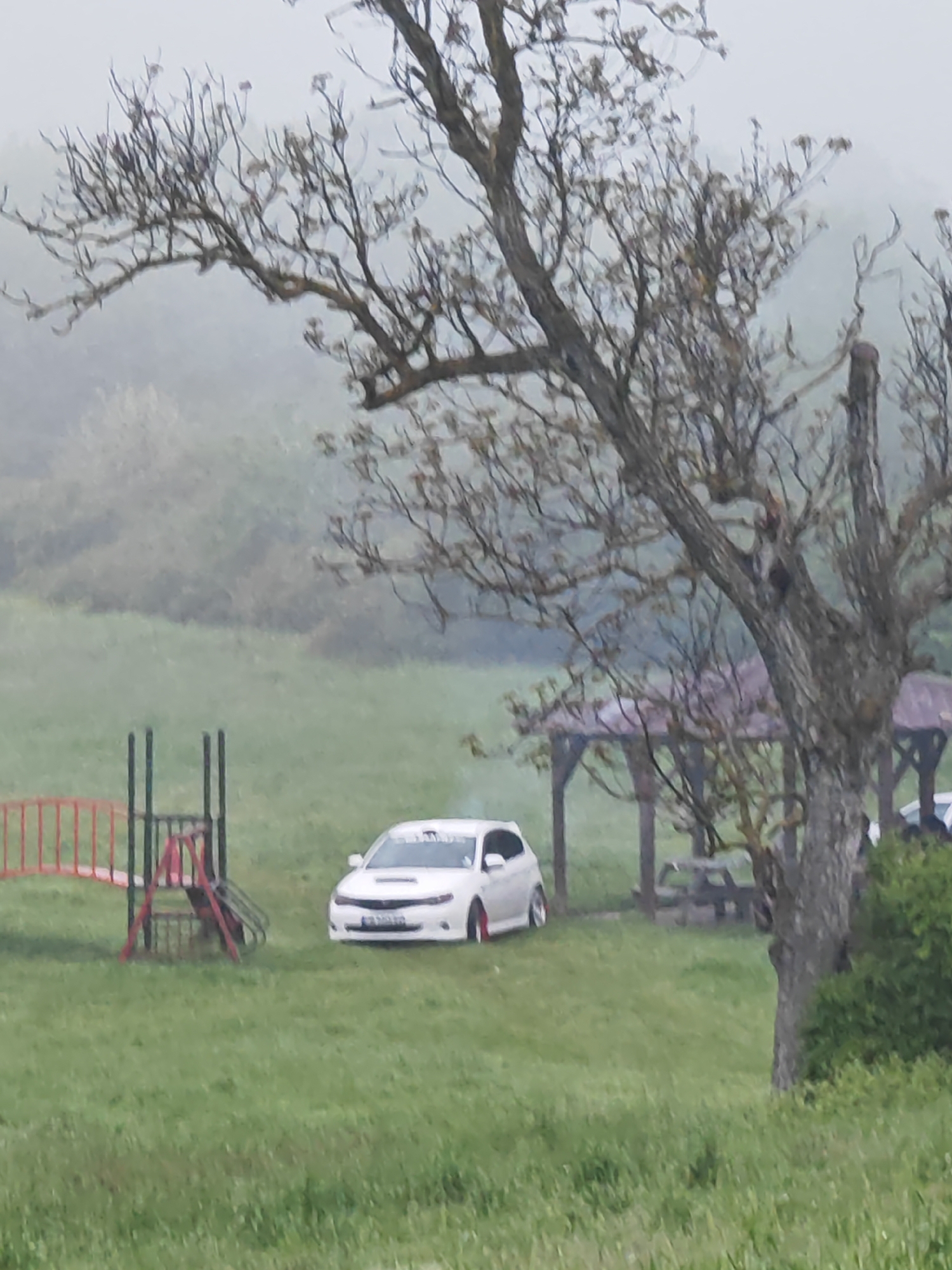 The image shows a grassy field with a white car parked near a playground and a gazebo. The environment appears misty or foggy, typical of a rural setting. The foreground is dominated by a tree with bare branches. There are no visible people. The location appears to be a park or recreational area. While I cannot definitively place it in Shumen, Bulgaria, based on the image alone, this setting could exist in the outskirts of the city or a nearby rural area.
