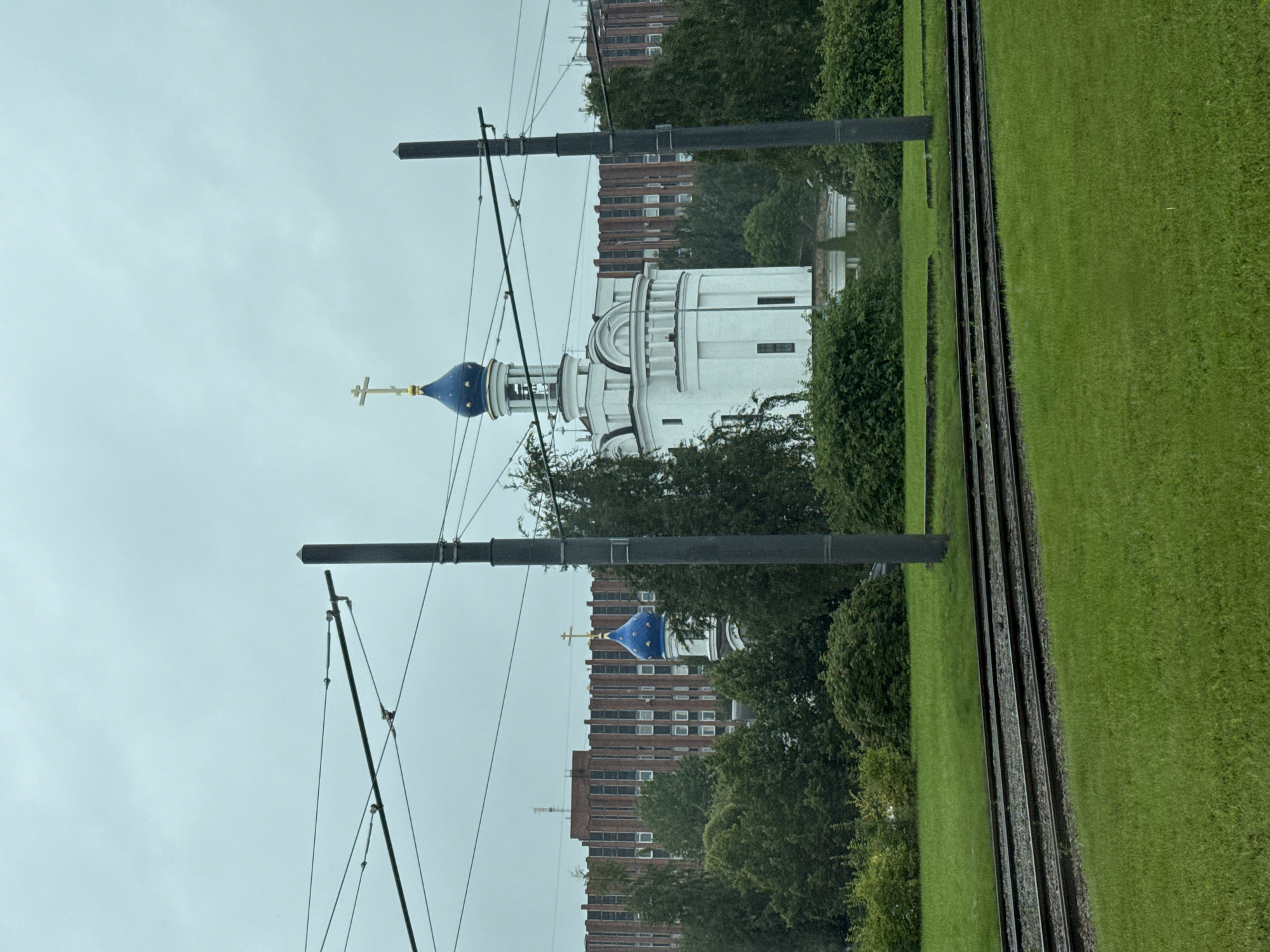 The image shows a bright white building with a blue domed roof topped with a golden cross, resembling a church. This building is situated amidst lush green trees and bushes. In the foreground, there are tram tracks laid on the ground, with overhead wires supported by poles, indicating an urban setting. In the background, a large, modern brick building with numerous windows stretches across the horizon. The sky is overcast and light blue, suggesting a cloudy day. The overall scene is a blend of religious architecture, urban infrastructure, and natural greenery. This scene is located in the city of Saint Petersburg, Russia, specifically in the area of Pushkin, near the Holy Trinity Cathedral and the Alexander Nevsky Lavra.