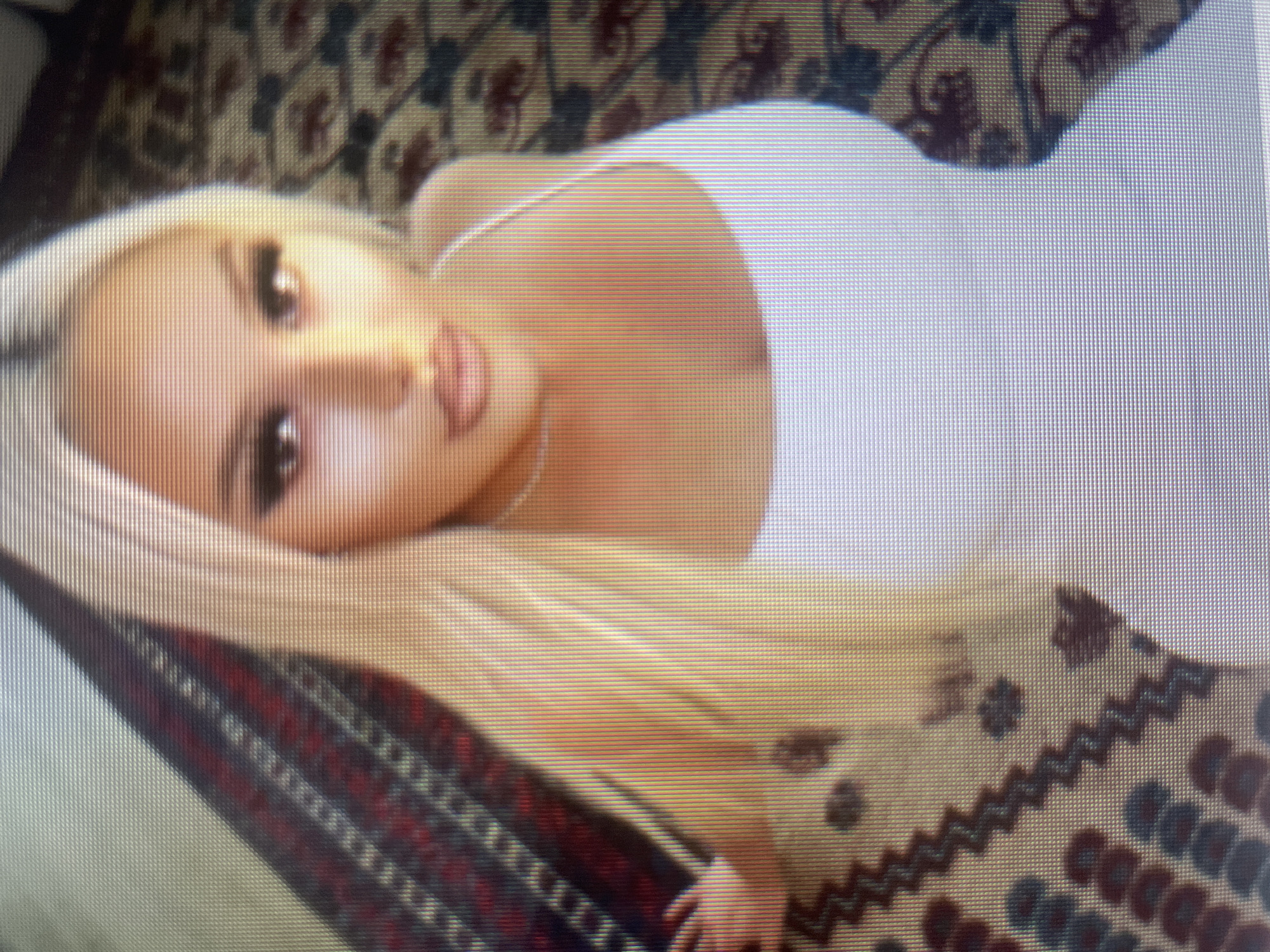 A woman with blonde hair and full lips, wearing a white tank top, is shown from a high angle, looking up at the camera. She is positioned on a rug with a patterned design. The image is pixelated, suggesting it was taken on a screen or with a low-resolution camera.