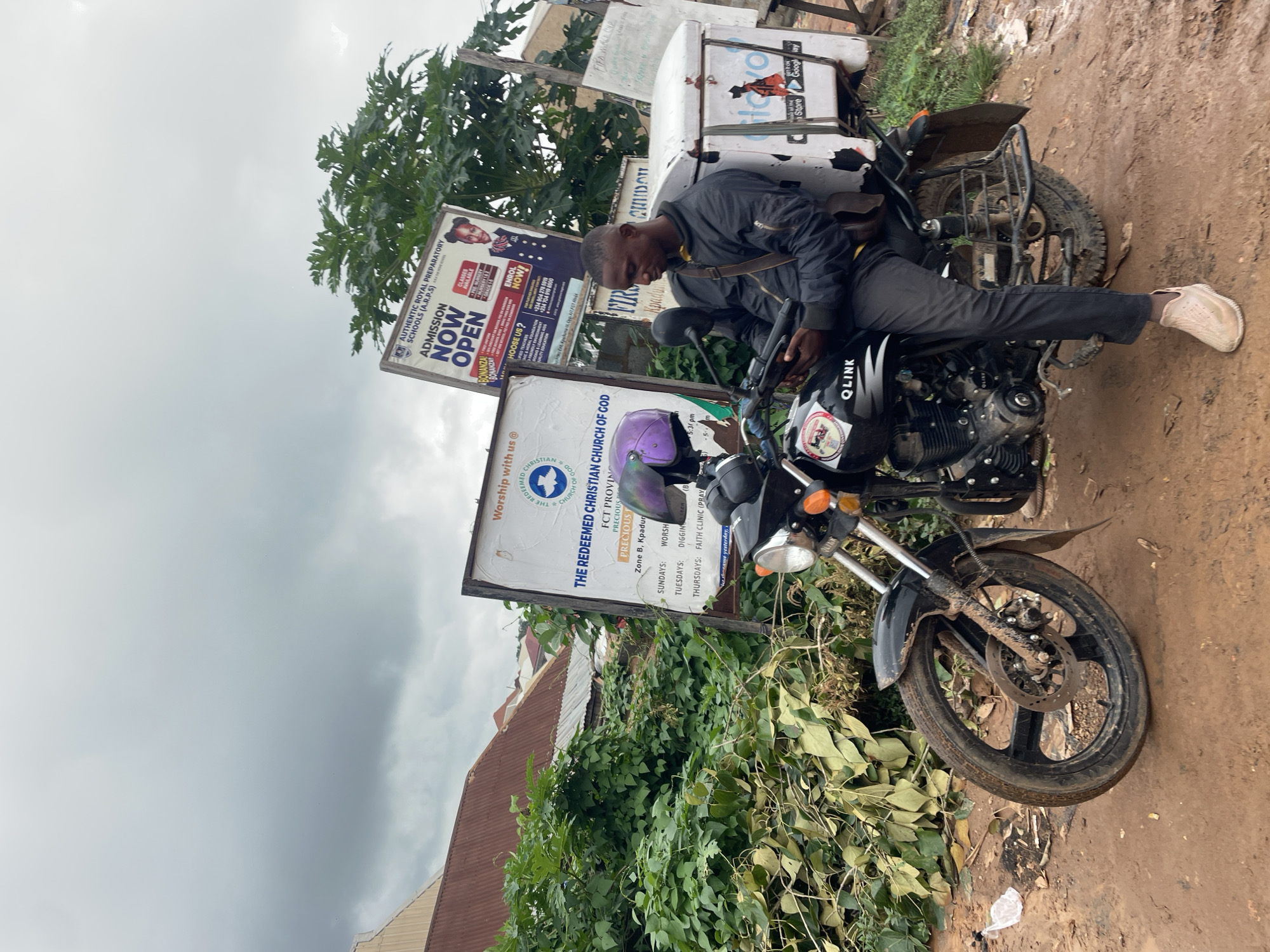 A Black man sits on a black motorcycle parked on a muddy dirt road in Kurunduma, Nigeria. He is wearing a black jacket, dark pants, and light-colored shoes. The man is looking down at a black electronic device held in his hands. The motorcycle has a white cargo box on the back with some writing on it and a purple helmet on the seat. Behind the motorcycle are several signs, including one that reads "THE REDEEMED CHRISTIAN CHURCH OF GOD" and another advertising "AUTHENTIC ROYAL PREPARATORY SCHOOLS (A.R.P.S) ADMISSION NOW OPEN". There is lush green foliage and some corrugated metal roofs of buildings visible in the background. The sky is overcast and appears to be cloudy.