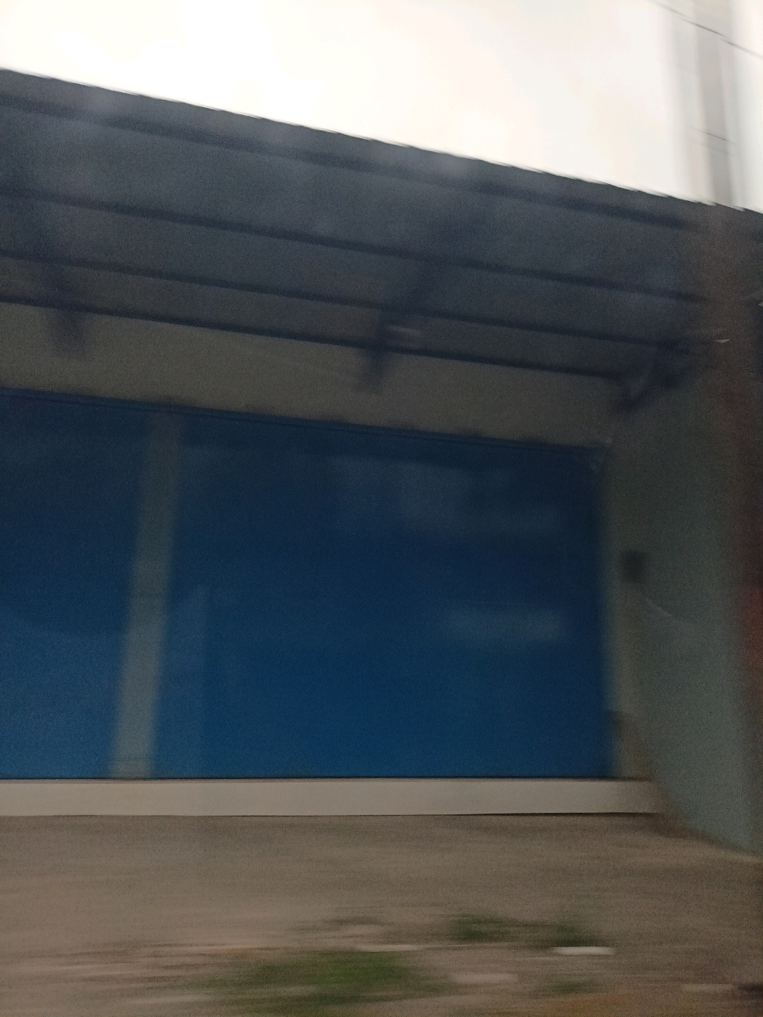 The image shows a closed building with large blue roller shutters. The building appears to be industrial or commercial, possibly a warehouse or a shop. The shutters are smooth and uniformly blue, with no visible markings or designs. Above the shutters, there is a dark overhang, possibly a roof or a canopy, supported by dark beams. The walls of the building are a light, neutral color, possibly concrete or painted. To the left of the shutters, there is a vertical white stripe that extends from the top of the shutters upwards. The foreground shows a blurry, grey surface, which could be a road or a pavement, with some faint green patches that might be grass or vegetation. The overall scene is somewhat out of focus, suggesting it was taken while in motion, possibly from a vehicle. The sky above is a pale, hazy white, indicating an overcast day. The image does not contain any people, animals, or specific landmarks that would help pinpoint a precise location within Gandusari, Indonesia, beyond the general context of a building in an urban or semi-urban environment.