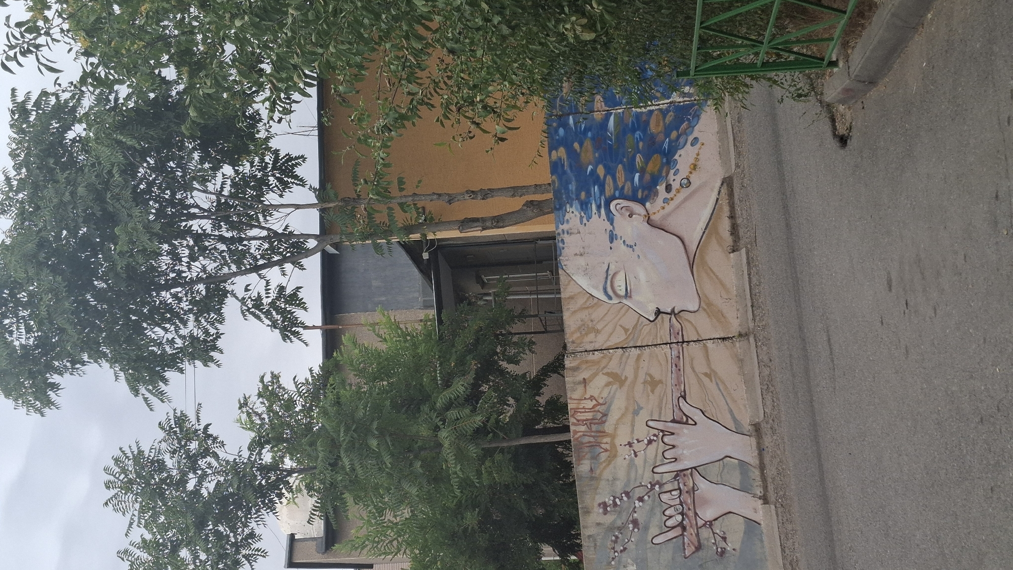 The image depicts a mural painted on a concrete wall in Shymkent, Kazakhstan. The mural features a stylized profile of a woman with long, blue, wavy hair adorned with golden droplets and beads. Her eyes are closed, and her mouth is slightly open, as if blowing. In front of her, a pair of hands plays a wooden flute. The background of the mural is a muted earth tone with faint depictions of birds in flight. Above the wall, trees with green foliage obscure parts of buildings, one of which has an orange facade. A gray and dark gray structure is also visible behind the trees. In the foreground, a paved road runs parallel to the wall. A green metal fence stands to the right of the mural, partially covered by vegetation. The sky is overcast.