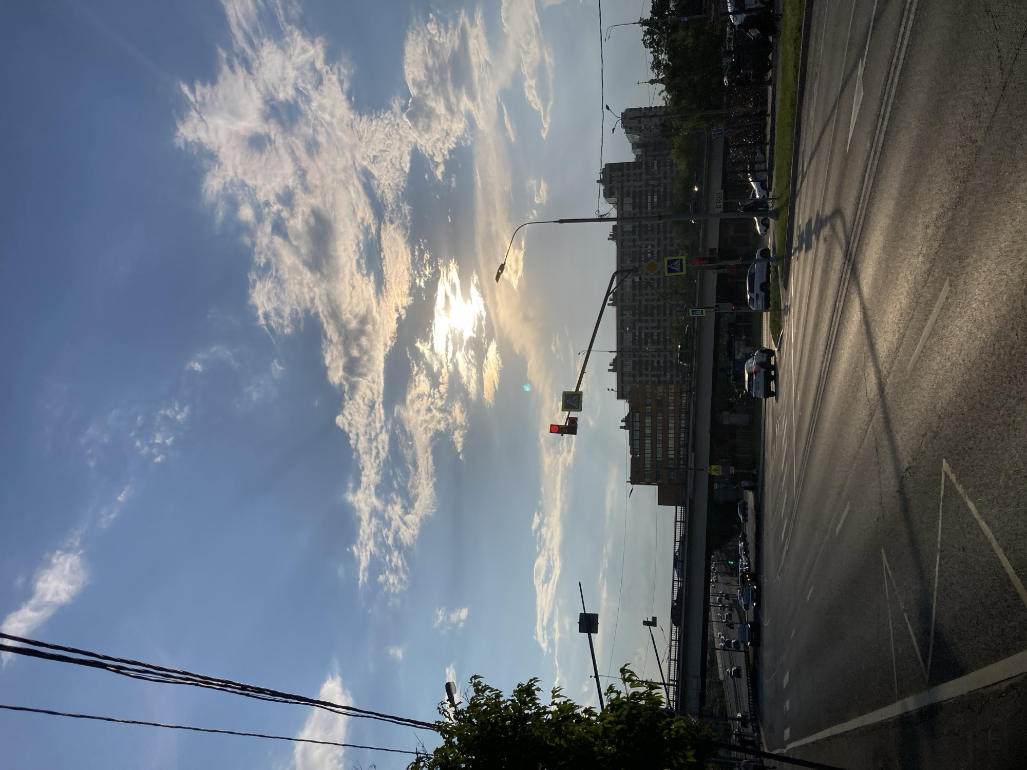 The image captures a wide-angle view of a street scene at an intersection in Moscow, Russia, likely during late afternoon or early evening, given the low angle of the sun and the shadows. The sky is a mix of blue with scattered white and grey clouds, partially obscuring a bright sun that casts rays through the cloud cover.
In the foreground, a multi-lane road is visible, with cars driving and stopped. Traffic lights are present, with one showing red. Pedestrian crossing signs are also visible. A street lamp stands on the right side of the road.
A bridge or overpass crosses the road in the mid-ground, with traffic visible on it. Behind the bridge, several large apartment buildings, typical of Soviet-era architecture, dominate the skyline. Some trees and greenery are present on the sides of the road and near the buildings. Overhead power lines stretch across the top left portion of the frame.
There are a few people visible on the sidewalk and at the pedestrian crossing. A person is walking on the left side of the road. Two people are standing near the sidewalk on the right side, and a person is riding a bicycle. Cars are the primary objects in motion. The overall scene depicts a busy urban environment with elements of infrastructure, residential buildings, and vehicular traffic.