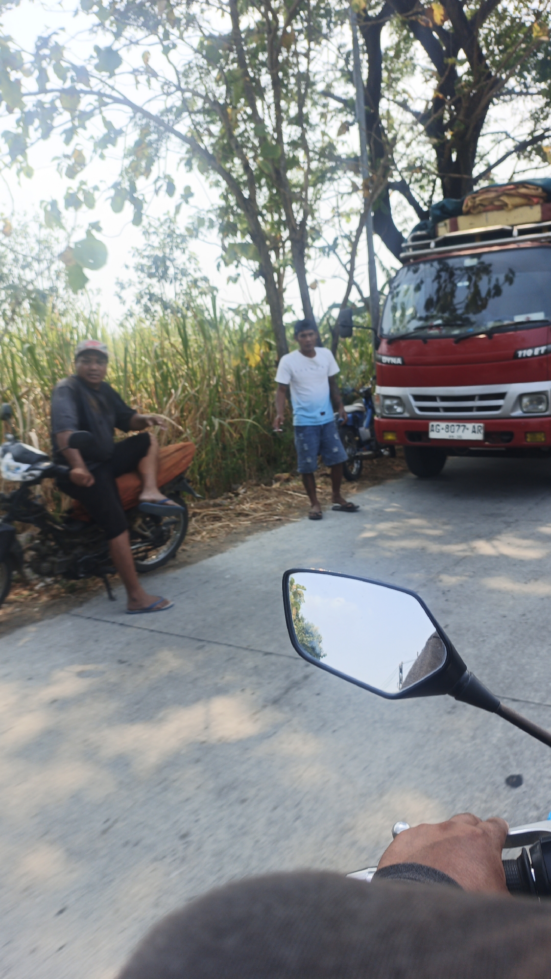 In Randugenengan, Indonesia, a scene unfolds on a paved road bordered by dry vegetation and tall plants. A red truck, identified by the license plate AG-8077-AR, is parked on the right side of the road. A man in a white t-shirt with a blue gradient and denim shorts stands to the left of the truck, holding a cigarette. On the left side of the road, a man wearing a dark t-shirt, a red and black cap, black shorts, and blue flip-flops is seated on a black motorcycle. The motorcycle has an orange seat cover and a white front. The perspective of the image is from the viewpoint of someone on a motorcycle, looking forward, with the motorcycle's rearview mirror reflecting the sky and trees.