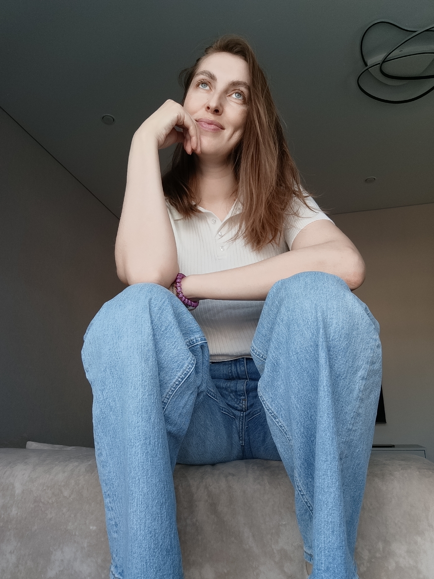 A young woman is featured in a low-angle indoor shot, seated with a thoughtful expression. She has light brown hair, blue eyes, and a serene, slightly smiling face with a visible dimple. She is wearing a light-colored, ribbed short-sleeve polo shirt and loose-fitting light blue jeans. Her left elbow rests on her knee, and her left hand supports her chin, while her right arm is also bent, resting on her other knee. A purple bracelet is visible on her left wrist.
She gazes upwards and slightly to her right, suggesting contemplation or looking at something out of frame. The setting appears to be a modern indoor space in Chaikovsky, Russia. She is sitting on a light grey, possibly upholstered surface, which could be a bed or a low sofa. The background consists of a plain grey wall and a ceiling where two light fixtures are visible: one recessed circular light and another decorative, black-outlined fixture with a curvilinear design. The lighting is soft and ambient, indicating it is likely daytime. There is no visible text in the image.