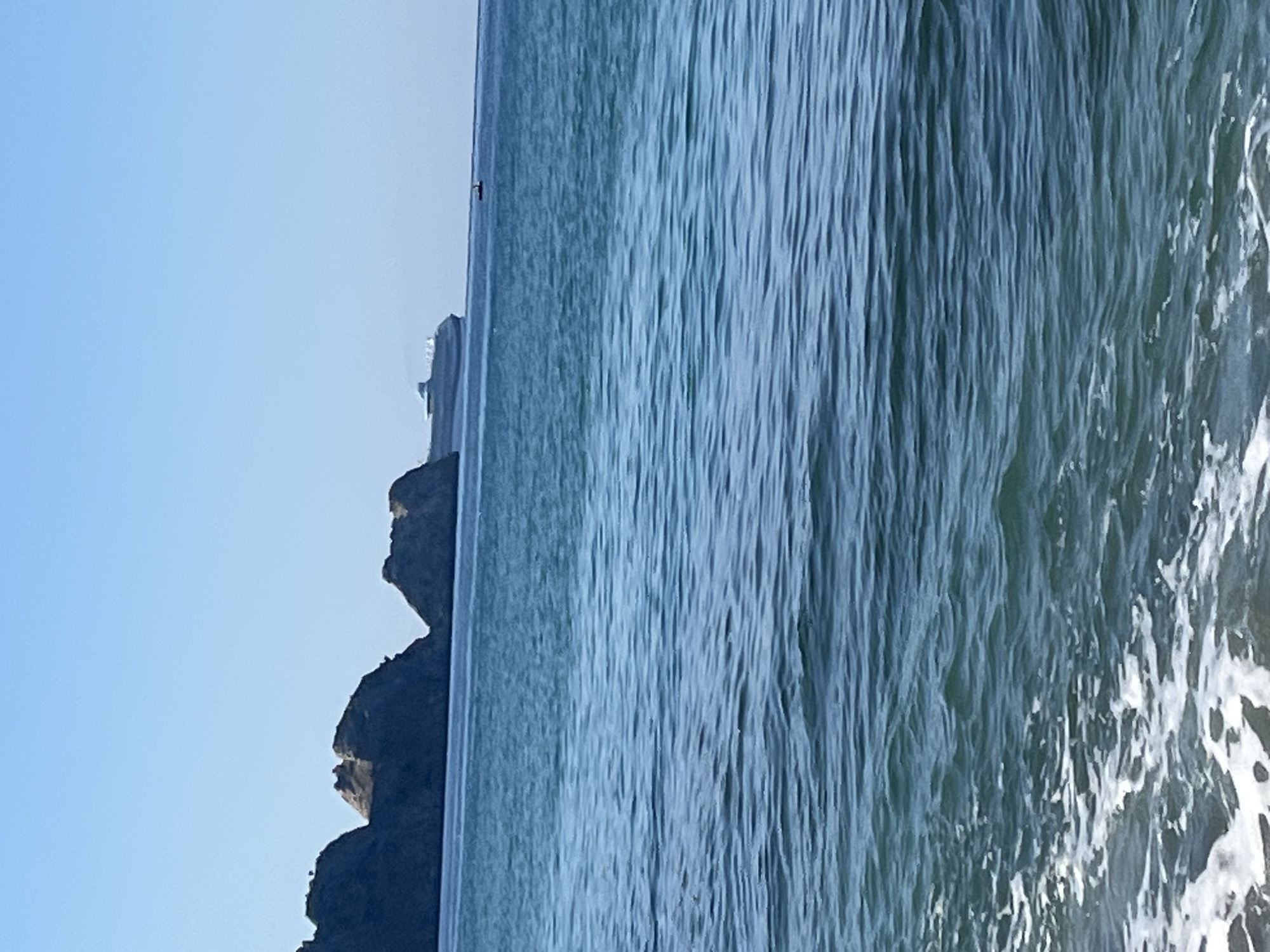 The image displays a serene coastal landscape, presenting a vast expanse of the sea under a clear, bright blue sky, bordered by dark, rugged cliffs. The sky, occupying the upper left portion of the frame, is a uniform, soft blue, indicative of a clear and sunny day.
The sea dominates the right side of the image, showcasing gentle waves and ripples that create a textured surface. The water exhibits varying shades of blue and green, with sunlit reflections adding brightness, particularly towards the foreground. Along the bottom-left, a series of dark, jagged rock formations rise, silhouetted against the bright sky, outlining a dramatic coastline or headland. Their irregular shapes suggest natural erosion.
On the distant horizon, a small, indistinct dark speck is visible where the sky meets the sea, possibly a distant boat or a small structure. The overall impression is one of tranquility and natural beauty. The bright, clear lighting suggests it is daytime, likely morning or early afternoon. This coastal scene is consistent with the provided location context of Alanya, Turkey. No text is visible in the image.