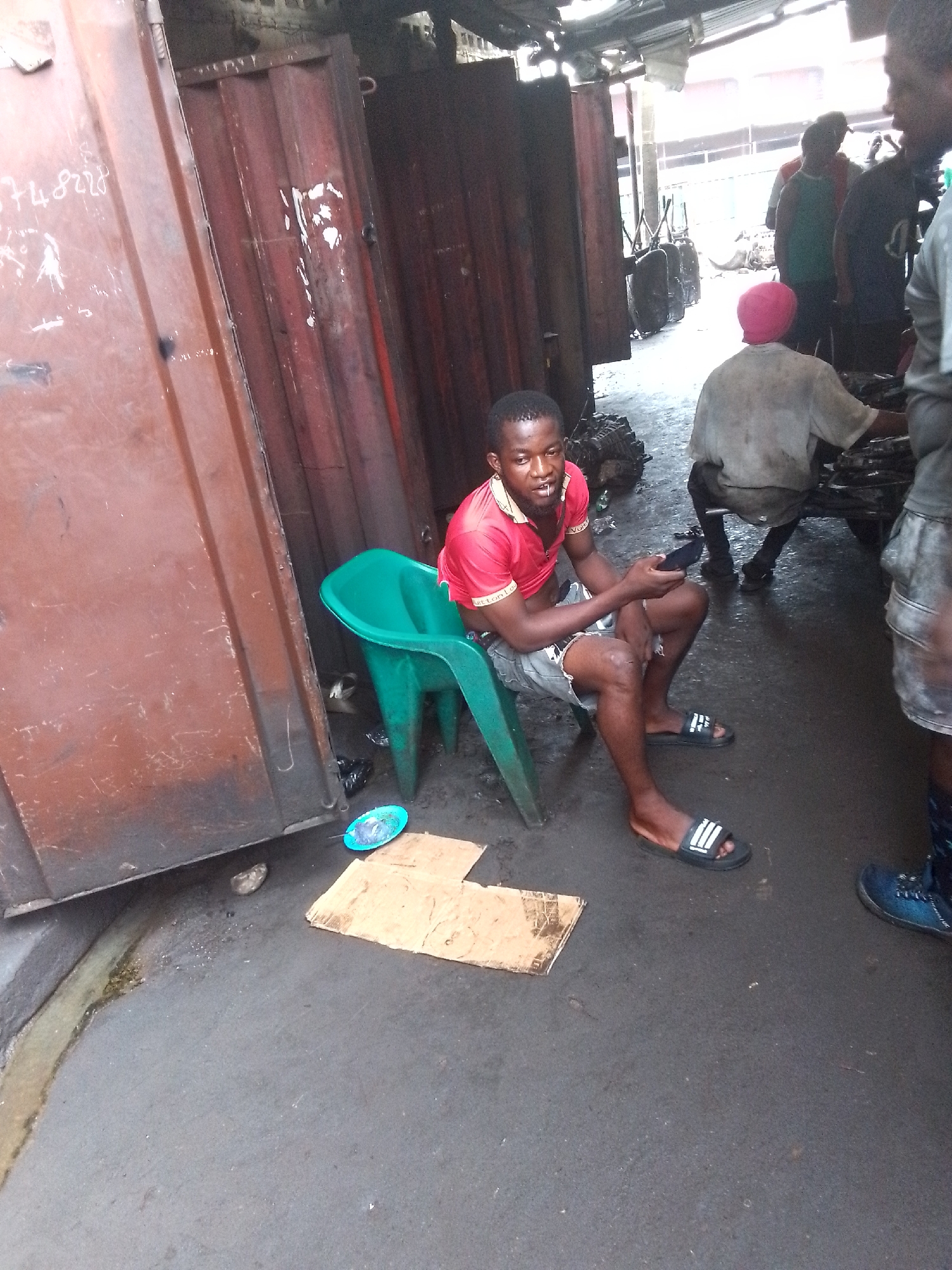 The image depicts a daytime scene in what appears to be an outdoor or semi-enclosed market or workshop area, likely in Onitsha, Nigeria.
In the foreground, a young man is seated on a bright green plastic chair. He is wearing a red polo shirt with faint branding, grey shorts, and black Adidas slide sandals. He holds a black smartphone in his hands and looks towards the camera, with a small white object, possibly a toothpick, in his mouth.
The immediate surroundings feature a dark, stained concrete or asphalt ground, with a small channel of liquid running along the base of a rusty brown corrugated metal wall, which dominates the left side of the frame. On the ground near the man, there are two pieces of flattened cardboard and a small, light blue/green plastic plate or bowl containing some indistinguishable items. A black plastic bag is also visible near the chair.
In the background, several other people are present, some partially obscured. One individual wears a pink head covering and a light-colored shirt, viewed from behind. Another person wears a green tank top. They are surrounded by various mechanical parts and debris scattered on the ground, suggesting an environment related to auto repair or spare parts. The overall scene conveys a busy, functional atmosphere.
Notable visible text includes white graffiti-like numbers "3748228" and other illegible markings on the rusty brown metal wall to the left. The Adidas logo with three stripes is clearly visible on the main subject's sandals.