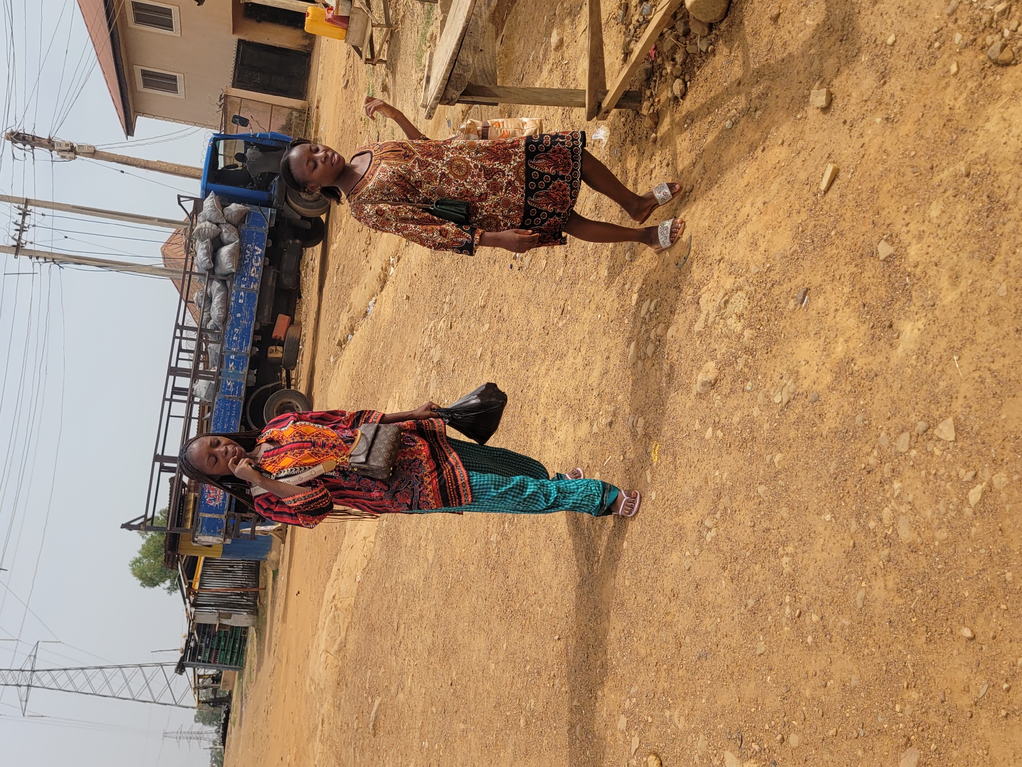The image depicts an outdoor scene in Jos, Nigeria, characterized by a reddish-brown dirt ground. Two women stand in the foreground. One is wearing a patterned dress, carrying a small purse. The other is wearing a colorful shirt, green pants, carrying a purse and a black plastic bag. Behind them, a blue truck loaded with bags (likely filled with charcoal or similar material) is visible. A building with a window and door is partially visible next to the truck. Electrical poles and power lines run along the left side of the frame, along with a power tower. In the background, shacks are erected alongside other buildings. A wooden table is to the right of the women.