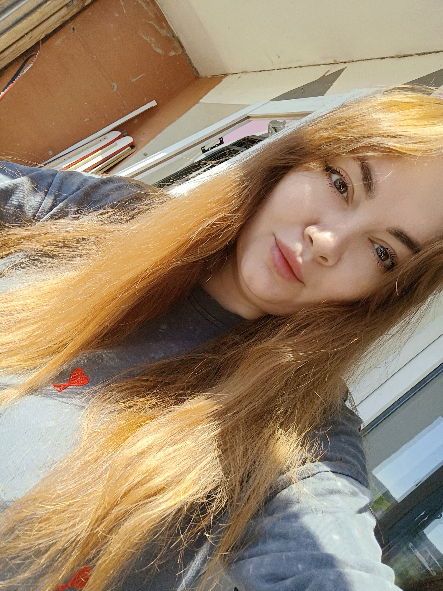 A woman with long, light-brown hair is looking at the camera. She is wearing a gray t-shirt with a small red heart on the chest. The location appears to be indoors, with a brown wall and a window in the background. The scene appears to be in Chaikovsky, Russia.