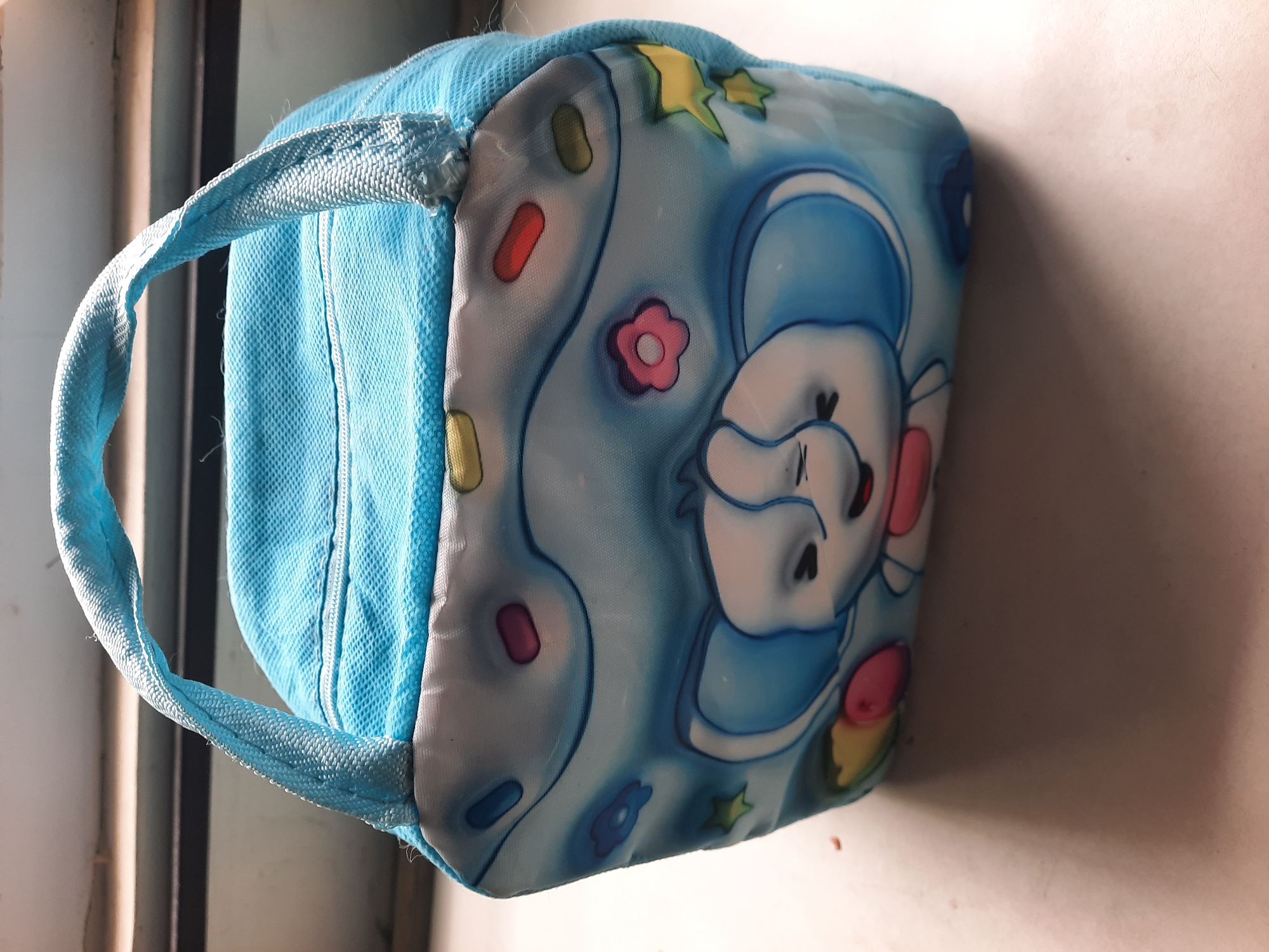 A blue fabric lunchbox with a cartoon elephant print sits on a white surface. The lunchbox has a zipper on top and a blue fabric handle. The elephant print is in shades of blue and white, with colorful accents. The background is blurred, but appears to be a window with light coming through. The lunchbox is located on a windowsill in Lhokseumawe, Indonesia.