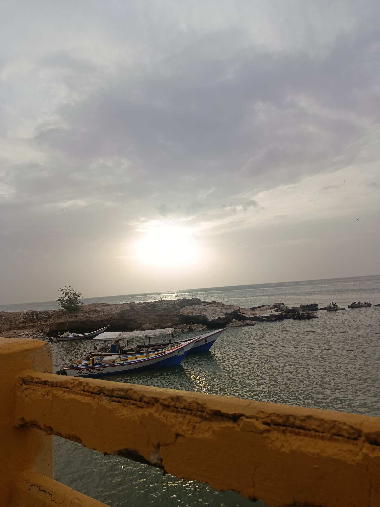 The image depicts a coastal scene in Juan Griego, Venezuela, taken on an overcast day with the sun trying to break through the clouds. In the foreground is a yellow concrete railing. Beyond the railing, there is a view of the water with several boats, one prominently labeled "La Patrona" in a white, red, and blue color scheme, docked near a rocky shoreline. Some of the rocks appear to be small islands, with dark shapes resembling birds perched on them. A small tree is visible on the main landmass. The sky is filled with gray clouds, with the sun creating a bright, diffused light over the water.
