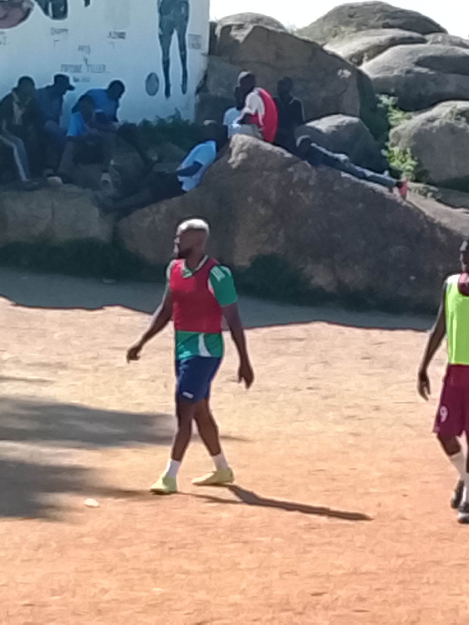 In Jos, Nigeria, a group of men are gathered near a wall with murals. Two individuals in the foreground appear to be involved in a soccer activity. One man, wearing a red vest over a green and white shirt and blue shorts, stands facing left. Another person, partially visible on the right, wears a lime green vest and burgundy shorts with the number "9" on them. In the background, several other men are seated on large rocks, some appearing to be observing the scene or resting. The ground is a dusty, reddish-brown surface, consistent with a dry, outdoor environment. The overall impression is a casual gathering, possibly related to a sporting event, in an outdoor urban setting.