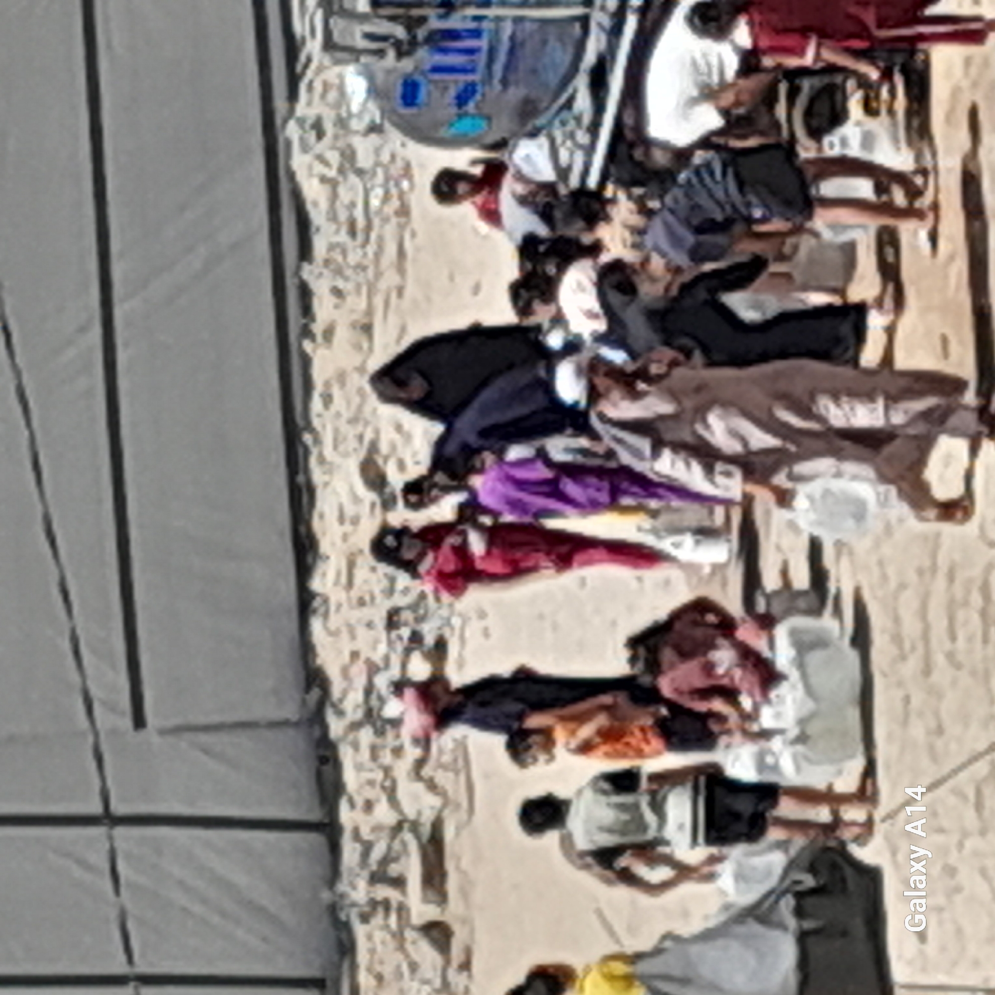 A group of people are gathered outdoors in what appears to be a dusty, unpaved area. The background is dominated by a large, gray, textured wall, possibly a building or barrier. In the foreground and midground, individuals are seen in various states of activity, many carrying containers, suggesting they are collecting or transporting water.
Several people are dressed in traditional clothing, including women wearing hijabs and abayas, and men in shirts and trousers. Some individuals are wearing brighter colored clothing, such as a red dress and a purple garment. There are children present, some of whom are also carrying containers.
In the right side of the image, a large, round, blue object, possibly a water tank or a vehicle, is visible. Some people are interacting with this object, and others are walking away from it. The ground is covered in sand and debris, and there are some makeshift shelters or tents visible, one of which is gray and black on the left side of the image. The overall impression is a scene of daily life, possibly involving resource collection or distribution in a somewhat austere environment. The watermark "Galaxy A14" is present in the bottom left corner of the image.