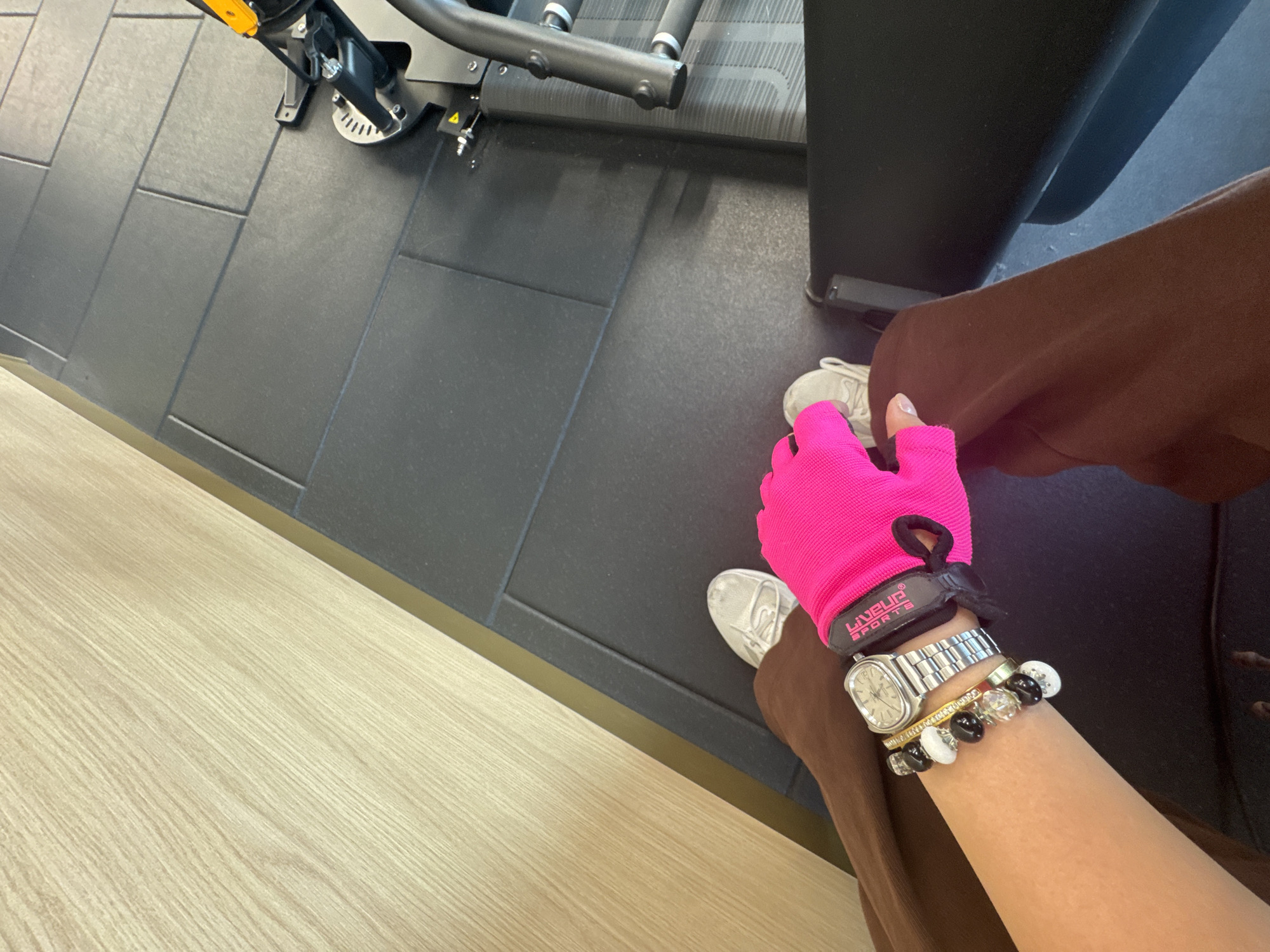 This image captures a first-person perspective within an indoor gym in Sofia, Bulgaria. The scene features the lower body and right arm of an individual, set against a dark grey tiled floor and a light wood-paneled wall separated by a thin gold strip. The individual wears brown athletic pants and white sneakers. The right arm, adorned with a silver-toned square-faced watch and multiple bracelets (a gold-toned bangle with small stones, and two beaded bracelets featuring black, white, and clear beads), extends into the frame. The hand wears a bright pink fingerless weightlifting glove with a black cuff displaying "LIVEUP SPORTS" in white text. Partially visible black and yellow gym equipment is situated on the right side of the frame.