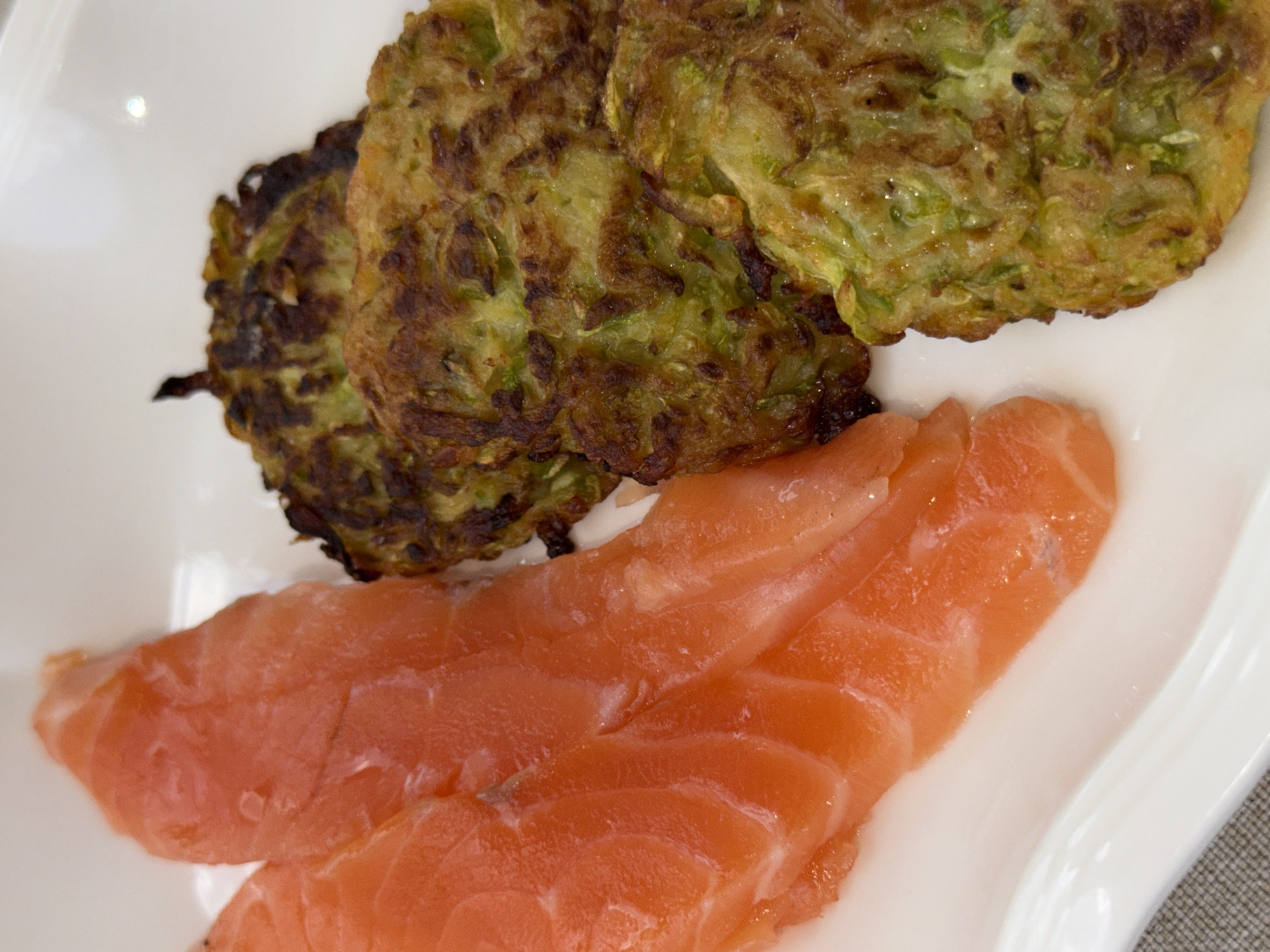 The image shows a close-up of a white plate on a textured gray surface. On the plate, there are several slices of raw salmon, which are a vibrant orange-pink color with white fatty streaks. Next to the salmon are several golden-brown, circular fritters, which appear to be made of grated vegetables, possibly zucchini or potato, with visible green flecks and darker, crispy edges. The food is arranged on the plate in a way that suggests it is ready to be served or eaten. The lighting highlights the textures and colors of the food, giving it a fresh and appetizing appearance. There are no people visible in the image, nor any indication of the specific location within Saint Petersburg, Russia.