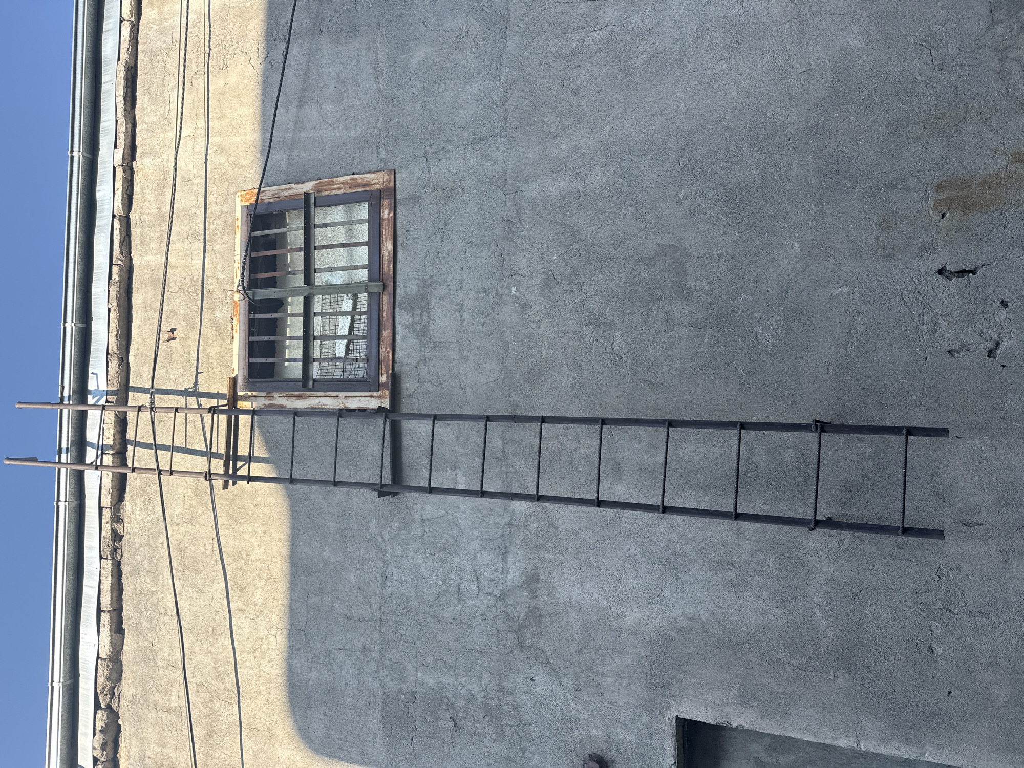 A metal ladder is attached to a rough, gray concrete wall of a building in Yerevan, Armenia. The ladder extends vertically along the wall, with multiple horizontal rungs. A small, barred window with a rusty frame is visible on the wall to the right of the ladder. Above the window and ladder, a corrugated metal gutter runs along the edge of the building, below a clear blue sky. The wall is partially illuminated by sunlight, casting shadows. To the left of the ladder, a portion of a dark doorway or opening is visible near the bottom of the frame. The concrete wall shows signs of weathering and cracking.