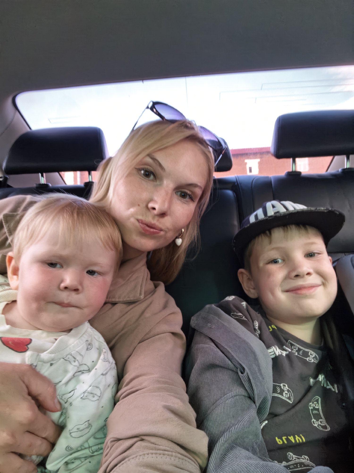 A woman and two children are inside a vehicle, possibly in the backseat, as indicated by the car's headrests and interior visible in the background. The woman, with blonde hair and wearing sunglasses on top of her head, is holding a baby on her left. The baby, with fair skin and light blonde hair, is looking towards the camera with a neutral expression. To the woman's right is a boy wearing a baseball cap and a t-shirt with a skateboard print. The boy is smiling and looking at the camera. The lighting suggests it is daytime. The environment visible through the car windows appears to be an urban setting with brick buildings. The image was taken in Sterlitamak, Russia.