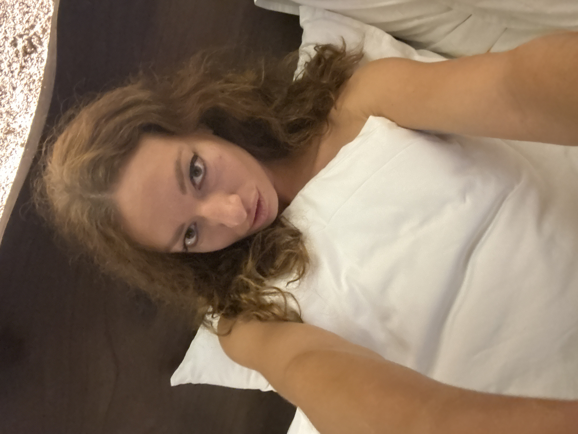 A young woman with long, curly brown hair lies in bed, propped up against a white pillow. She is looking directly at the camera, her expression neutral. Her left arm is visible, resting on the pillow. The background is dark, with a textured surface visible at the top of the image. The location is not specified in the image, and therefore cannot be determined to be Nam Hoa Lư Ward, Vietnam.