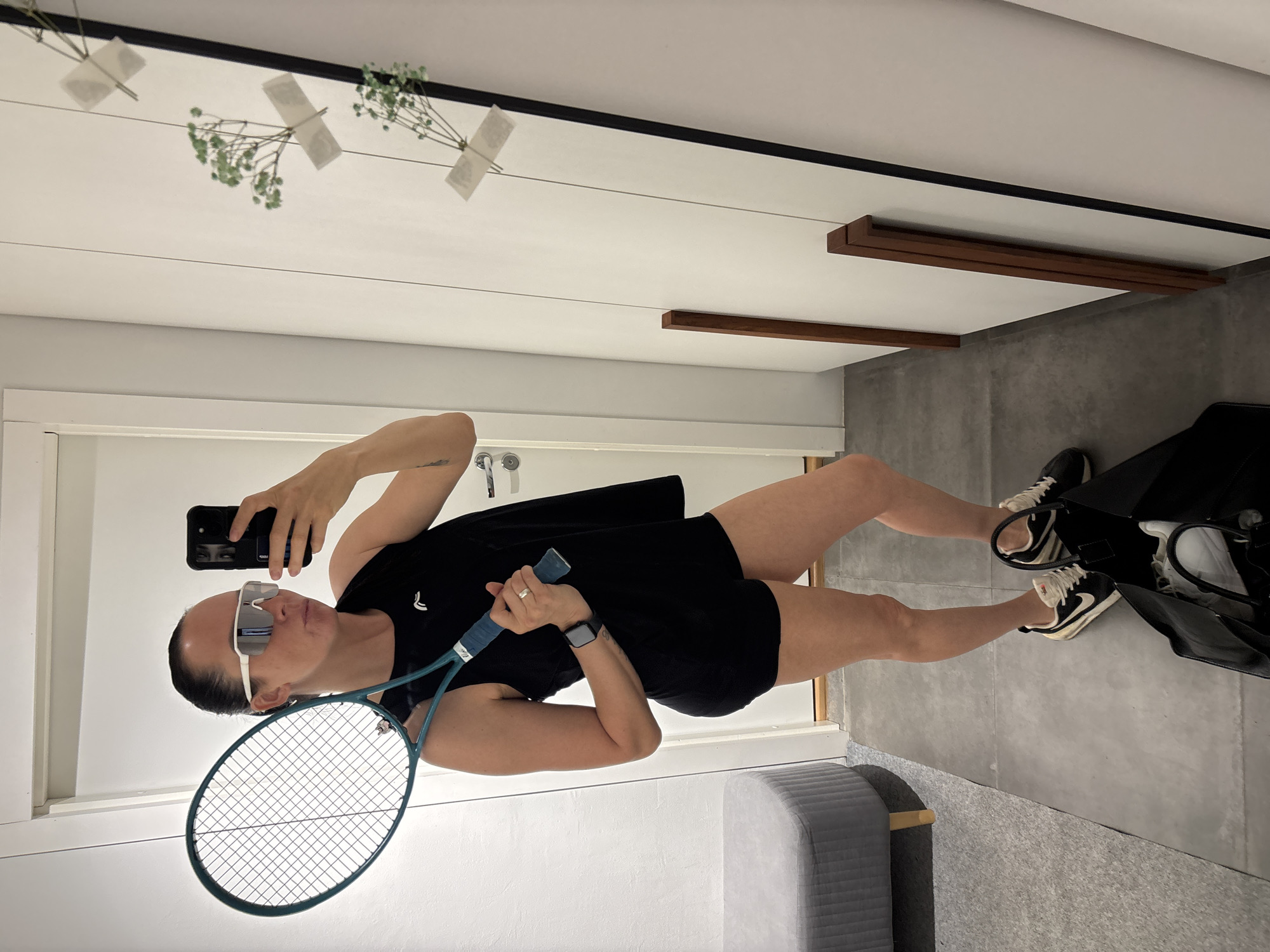 A woman stands in a brightly lit indoor space, possibly a home, holding a tennis racket and a smartphone. She wears sunglasses, a black athletic top, and black shorts. Her shoes are black and white sneakers. She is positioned on a gray floor with a light gray tufted ottoman behind her. A black bag rests on the floor. Behind her is a white door with silver hardware. The ceiling is white with decorative wooden elements. The image appears to be taken as a selfie. The location is unknown and cannot be determined to be the city of Chaikovsky, Russia.