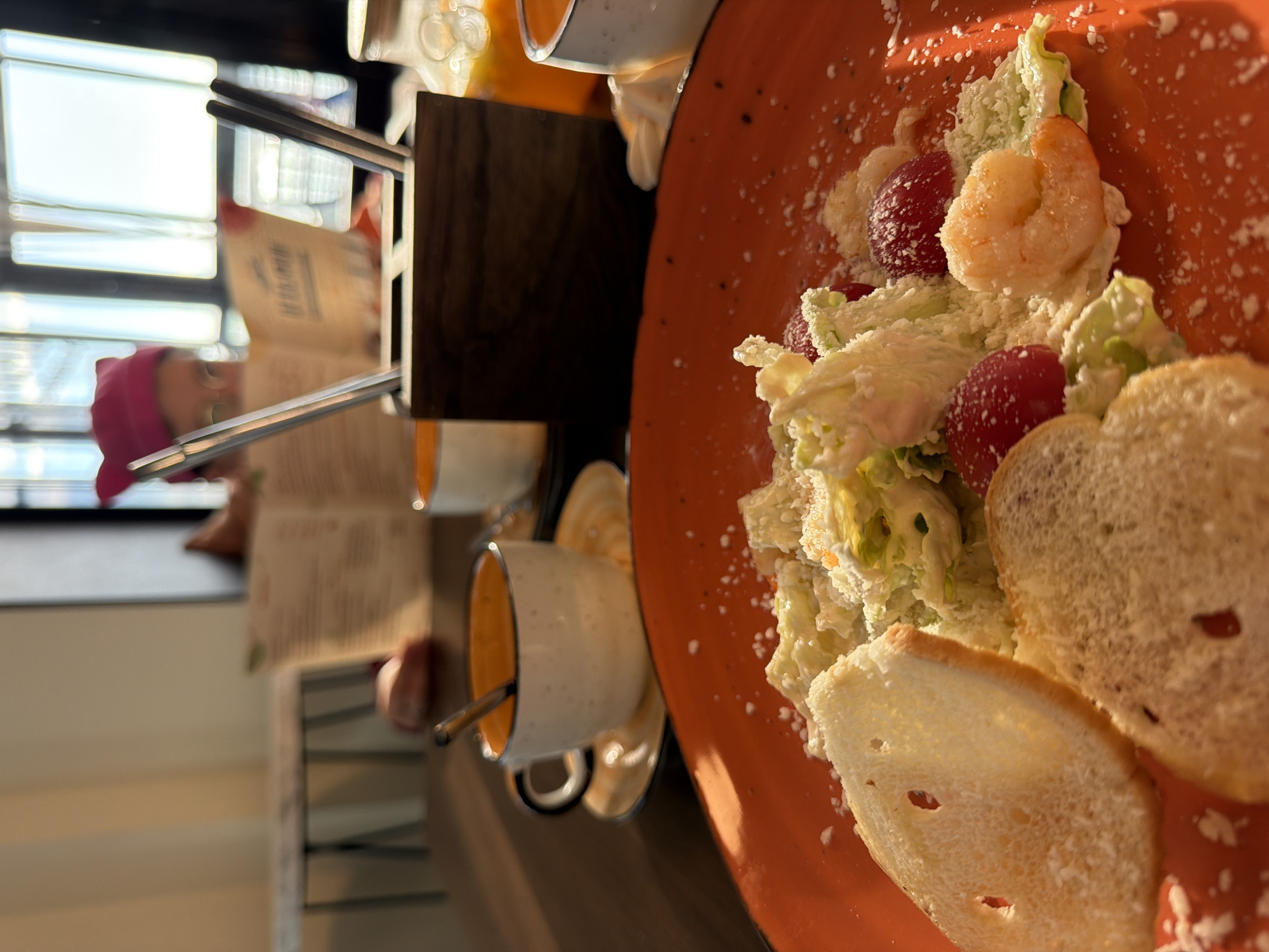 The image, taken in Chaikovsky, Russia, captures a dining scene in what appears to be a cafe or restaurant during daytime, illuminated by bright sunlight streaming through a large window. The composition is at an oblique angle, with the table and its contents appearing rotated.
In the foreground, a vibrant orange plate holds a fresh salad. The salad features crisp green lettuce, several bright red cherry tomatoes, and at least two plump, cooked pinkish-orange shrimp. The ingredients are coated in a creamy white dressing and generously sprinkled with grated white cheese or parmesan, along with specks of black pepper. Two slices of lightly toasted white bread or croutons are positioned at the bottom right of the plate.
In the blurred background, a person is partially visible, wearing a pink or magenta head covering. They are seated at a table and appear to be engrossed in reading an open book or menu, though the text on its pages is illegible. On the wooden table, next to the food, are two ceramic cups with white exteriors and contrasting orange interiors, each with a spoon resting inside. The bright light from the window suggests a sunny day.