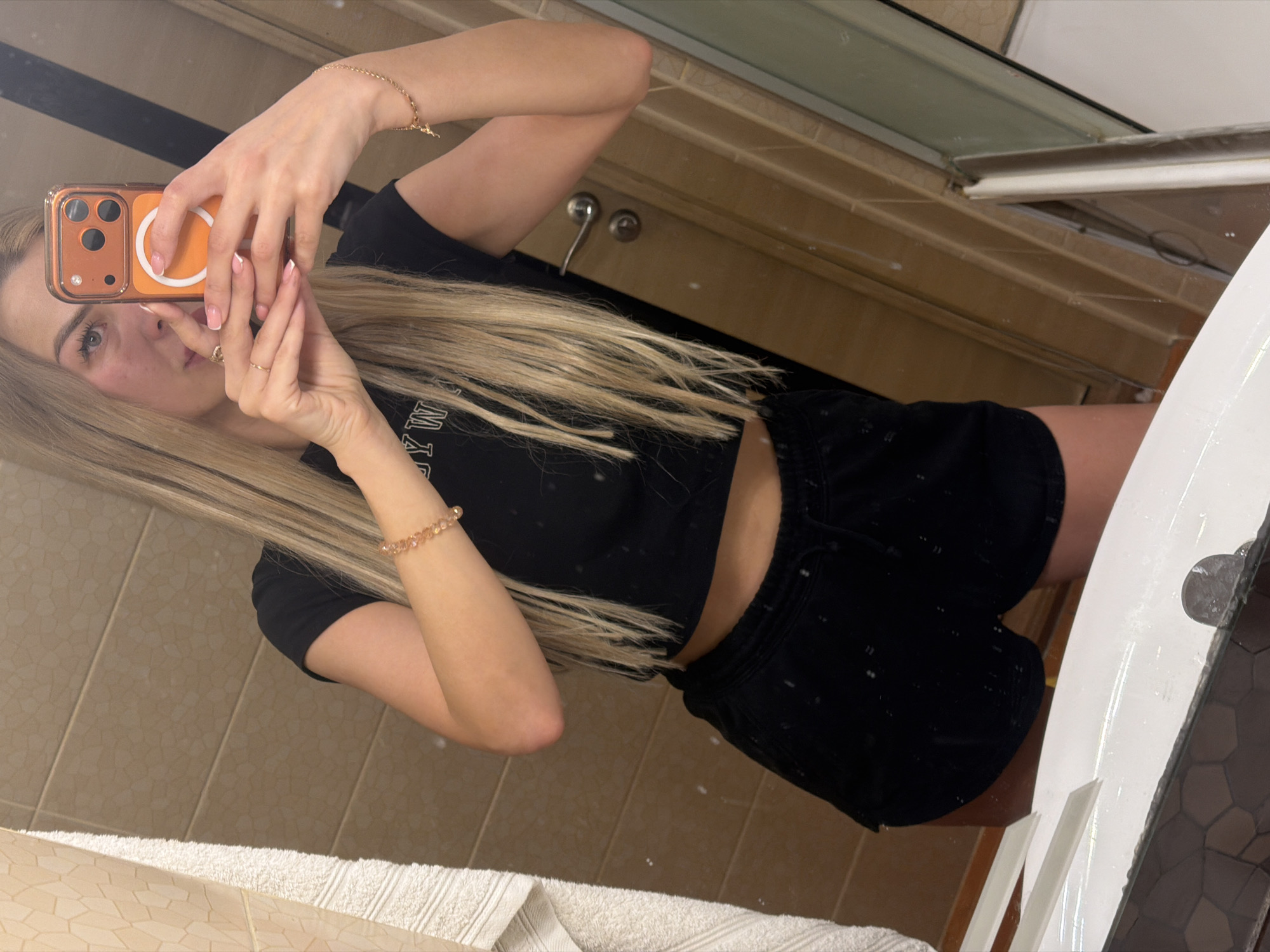 A young woman with blonde hair, wearing a black crop top and black shorts, takes a selfie in a bathroom. She is holding an orange phone in her right hand and a bracelet is visible on her left wrist. The bathroom has tiled walls and a mirror. The photo is taken from the waist up, and the lower half of her body is visible. The scene appears to be a private residence in Kyiv, Ukraine.