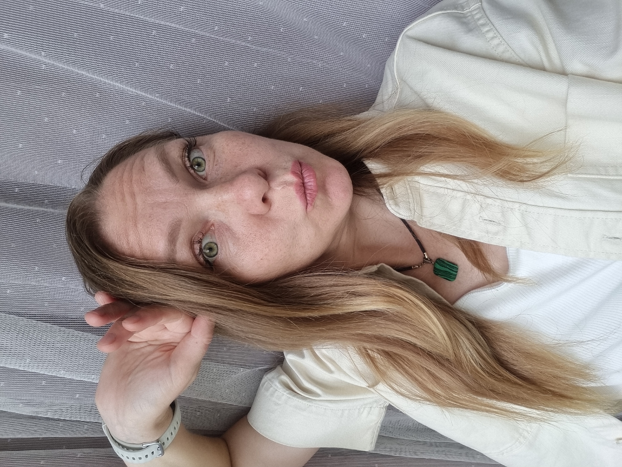 A medium shot, eye-level, indoor selfie shows a Caucasian woman with long, light brown hair, pale skin with freckles, green eyes, and light pink lips pursed. She is wearing a white tank top and a cream-colored collared jacket. A dark cord necklace with a rectangular green pendant is visible at her throat. Her left arm is raised, with her hand resting on her forehead and side of her head. She is wearing a white digital watch on her left wrist. The background consists of a vertical striped curtain in muted gray and white tones, appearing to be an interior setting in the city of Chaikovsky, Russia.