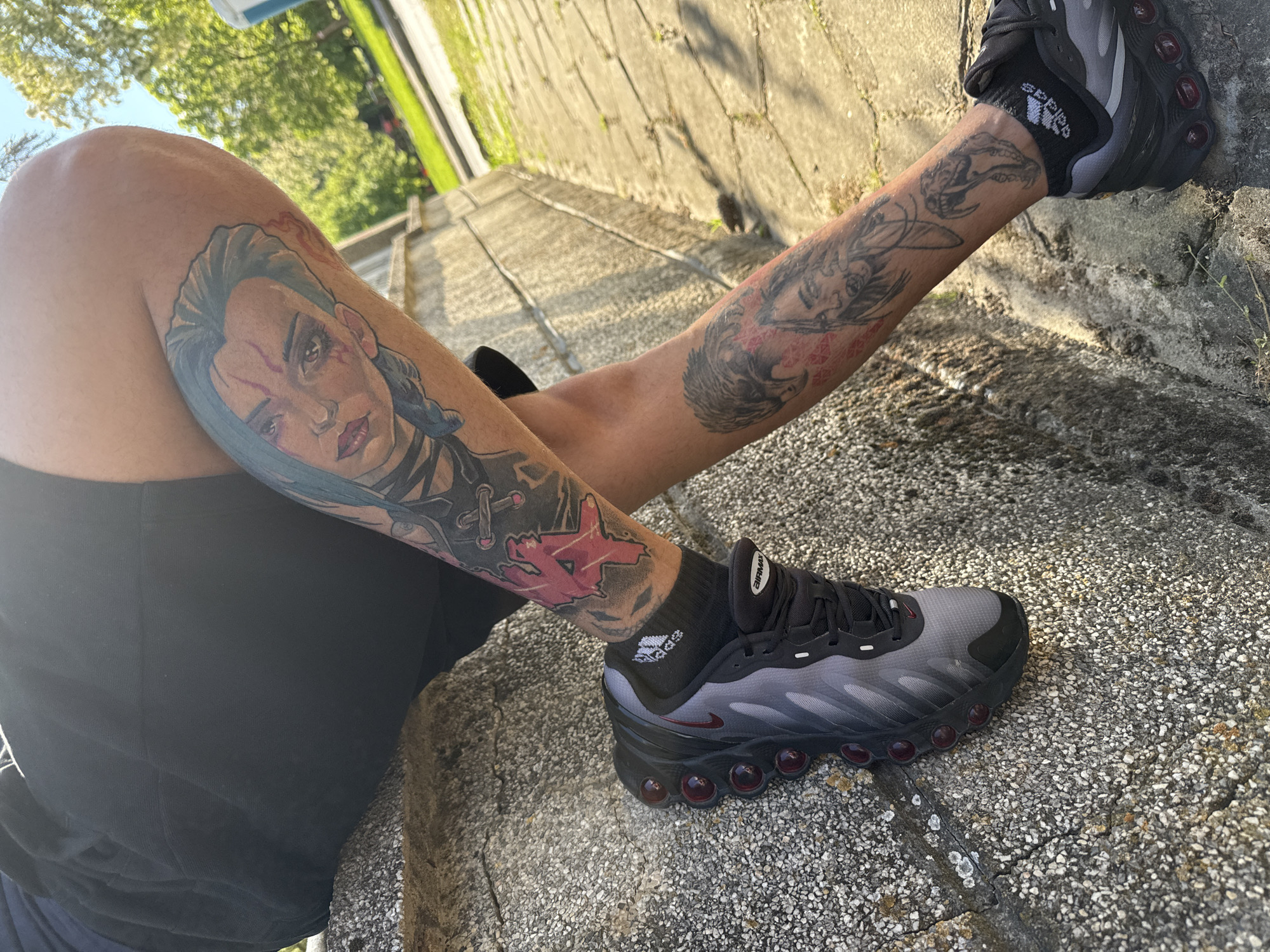 The image shows a person sitting outdoors. They are wearing black shorts and Adidas socks with black and gray running shoes. Their legs are extended, with one leg resting on a concrete ledge or step. Both legs have prominent tattoos. One leg features a tattoo of a woman with blue hair and a stern expression. The other leg has a tattoo of a stylized bird and other designs. The background appears to be a stone or concrete wall and some green foliage. The location is unspecified.
