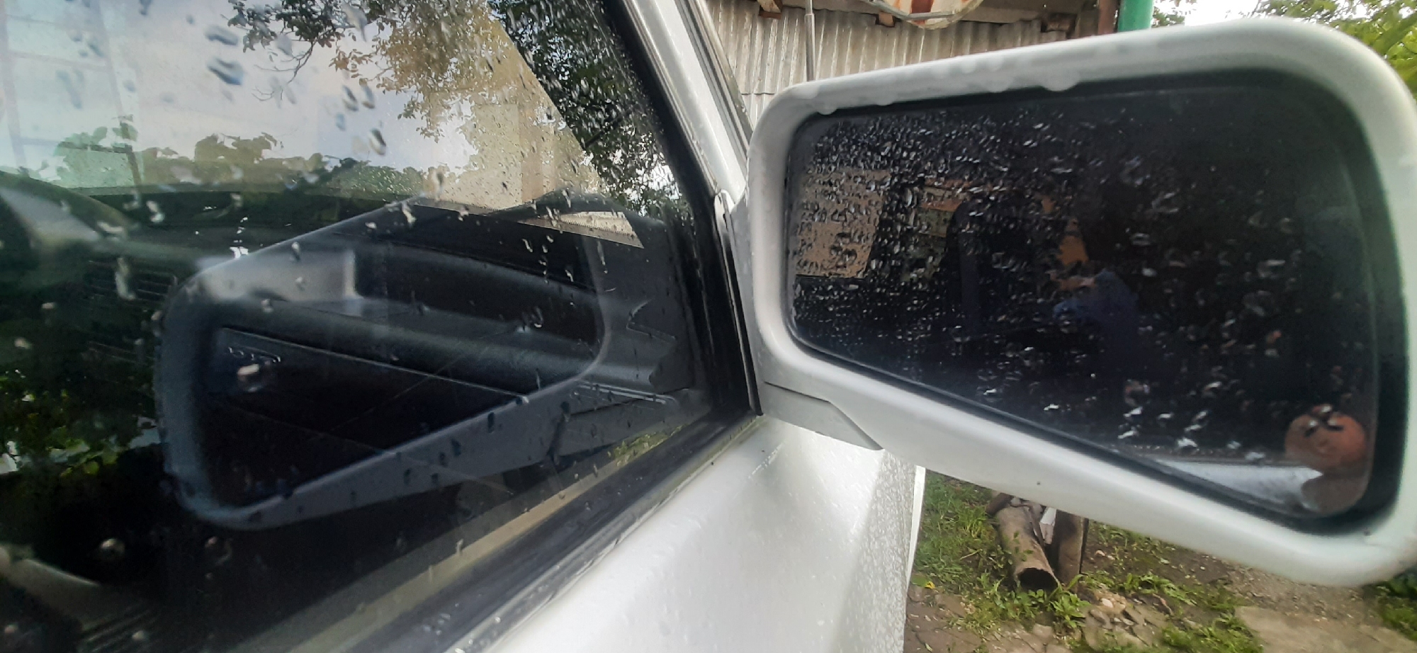 The image captures a close-up view of a white vehicle's side mirror and window, both of which are wet with raindrops, suggesting it might be raining or has recently rained. The side mirror housing is white and appears slightly aged, with water droplets distorting the reflection. The reflection in the mirror shows a person with dark hair wearing a blue shirt, their face partially obscured by water streaks. In the background, the reflection also hints at a wooden structure and possibly some greenery.
The vehicle's window, also streaked with raindrops, reveals the interior. A black plastic panel, likely part of the door or dashboard, is visible, with what appears to be a speaker grille. The reflection in the window shows trees and a building with a corrugated metal roof.
The overall scene is outdoors, with the ground visible at the bottom of the frame consisting of dirt and some green vegetation, including what looks like a fallen log. The lighting suggests it is daytime, but overcast due to the rain. The presence of a log and the rustic building in the reflection suggests a location that is not in a densely populated urban area, but could be in a suburban or rural setting. The mention of Yessentukskaya, Russia, places this scene in a specific geographic context.