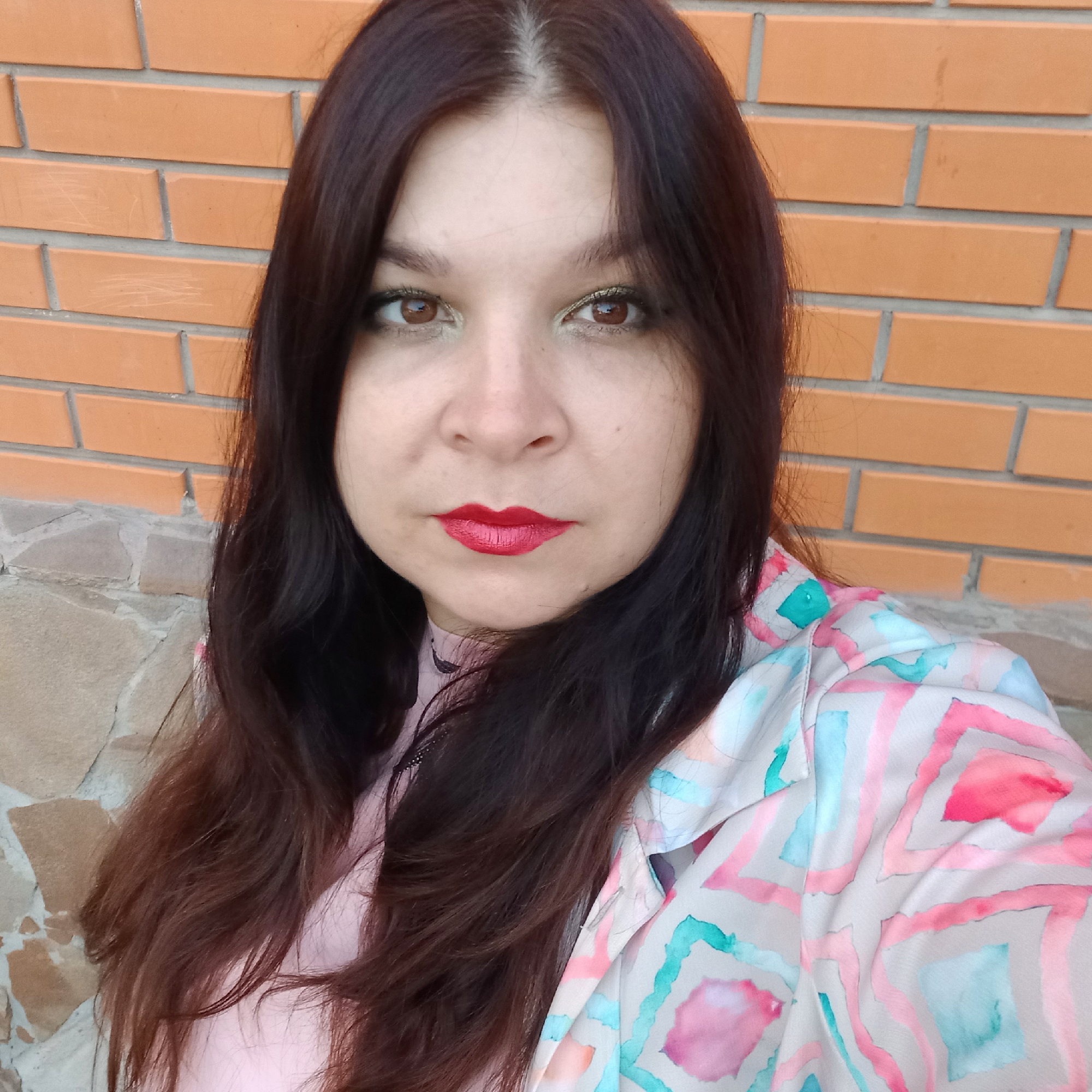 A young woman with long, dark brown hair and light skin is pictured outdoors, positioned in front of an orange brick wall. She is looking directly at the camera, her head slightly tilted. Her eyes are brown with noticeable golden eyeshadow, and she is wearing bright red lipstick. Her clothing appears to be a light pink shirt underneath a light-colored jacket with a pastel geometric pattern in shades of pink, blue, and yellow. The background consists of the brick wall and some rough-hewn stone at the bottom left. The image quality is somewhat grainy and exhibits a warm color cast. No specific landmarks or features are visible to pinpoint a location within Kropyvnytskyi, Ukraine.