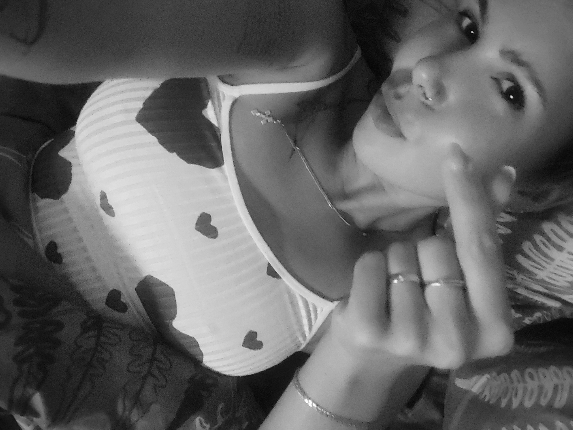 A monochrome image shows a woman lying down. She has a nose ring and is wearing a tank top with hearts and stripes. She is wearing a necklace and rings. Her right hand is raised towards her face. The background includes patterned fabric. The location is specified as Chaikovsky, Russia.