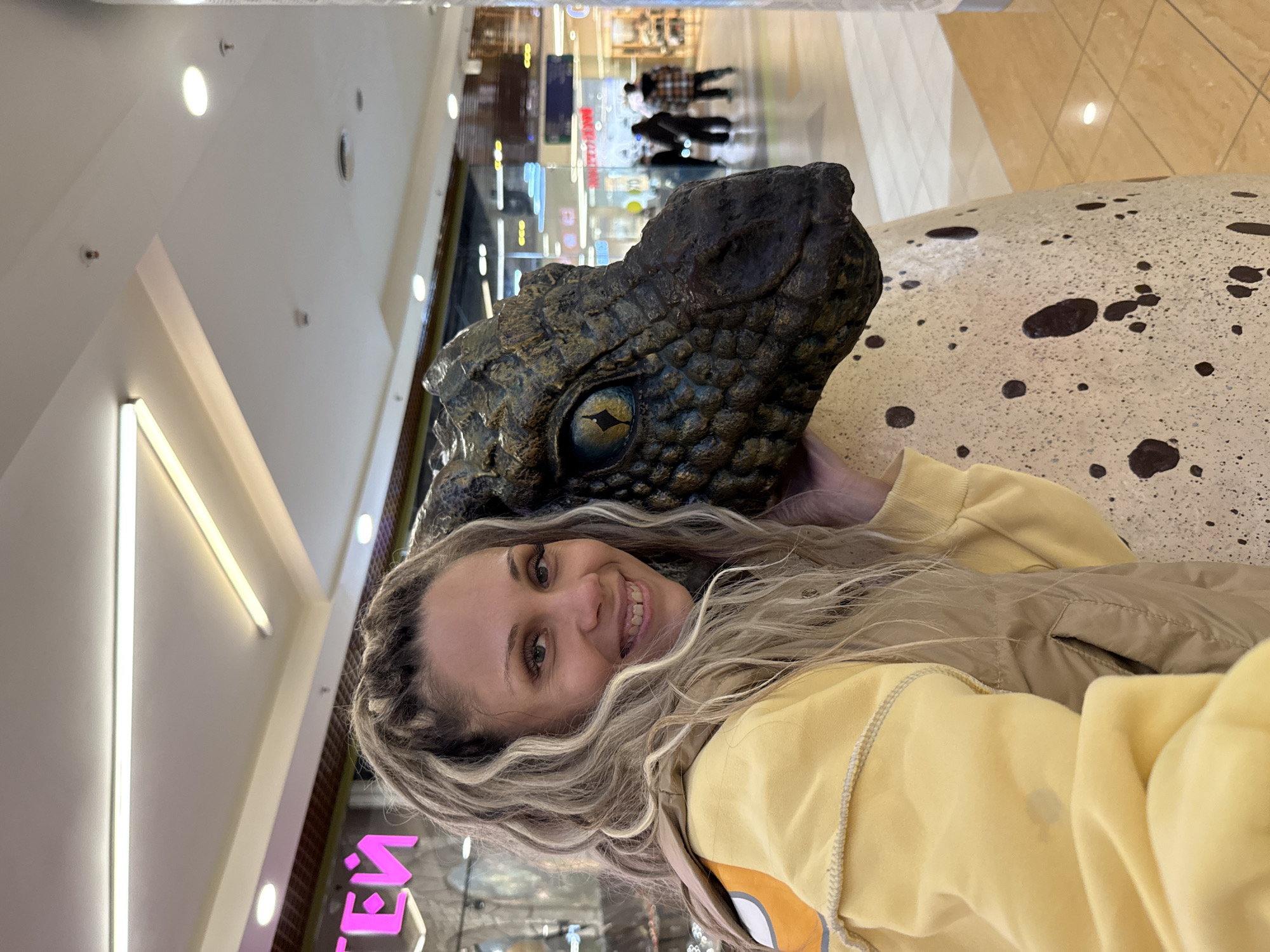 A woman with blonde hair styled in braids smiles at the camera, resting her head against a large, speckled egg with a reptilian head emerging from it. The woman wears a yellow hooded sweatshirt and a beige vest. The background appears to be an indoor shopping mall, with shops and people visible in the distance. The overall lighting suggests an indoor environment, possibly during the day. The woman's pose and proximity to the egg-like structure suggest she is interacting with a decorative display or sculpture. The scene is brightly lit, with fluorescent lights visible on the ceiling.