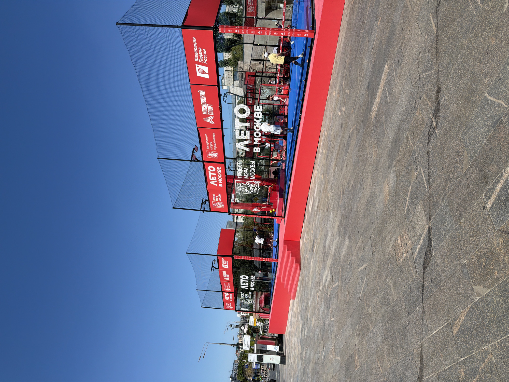 The image depicts an outdoor padel tennis court set up in a public space in Moscow, Russia, under a clear blue sky. The court is enclosed by black netting and features bright red barriers and a blue playing surface. Several individuals are visible on and around the court, some playing padel tennis and others observing. Large banners with Russian text and logos are displayed on the enclosure, including "ЛЕТО В МОСКВЕ" (Summer in Moscow), "ПРОЕКТ МЭРА МОСКВЫ" (Moscow Mayor's Project), "МОСКОВСКИЙ СПОРТ" (Moscow Sport), and "Федерация Падела России" (Padel Federation of Russia). In the background, city buildings and streetlights suggest an urban environment. The ground in front of the court is paved with large, grey stone tiles.