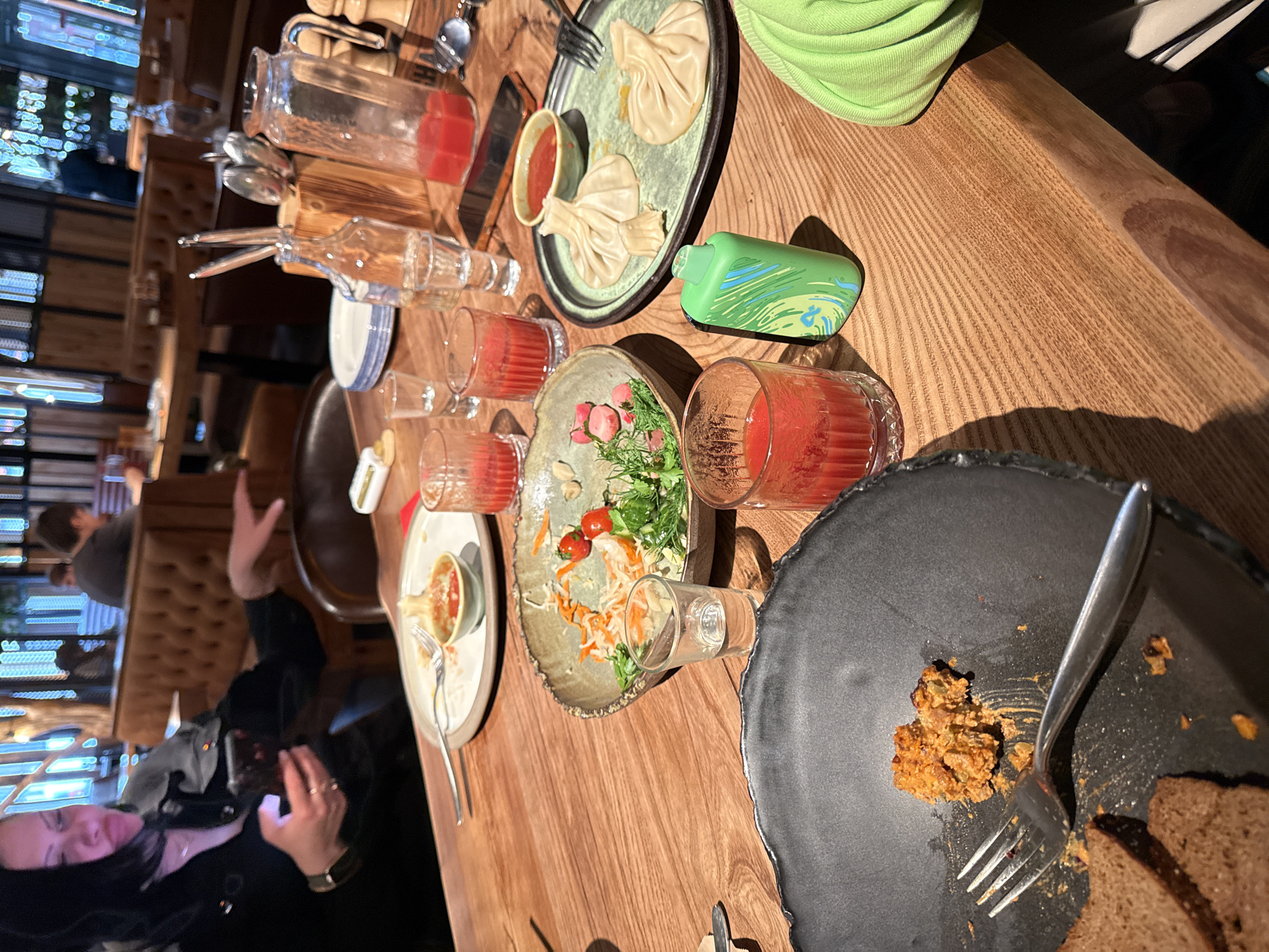 The image depicts a table setting in a restaurant, potentially located in Malakhovka, Russia, although this cannot be definitively confirmed from the image alone. The table, made of wood, is covered with various dishes and glasses. There are plates containing partially eaten food, including what appears to be salad, dumplings, and bread with some type of spread. There are also several glasses and a pitcher containing a red liquid, possibly juice or a cocktail. Cutlery is scattered around the plates.
A person is visible in the lower left of the image, partially obscured, holding a phone. The location and details of the city of Malakhovka cannot be verified from the image.