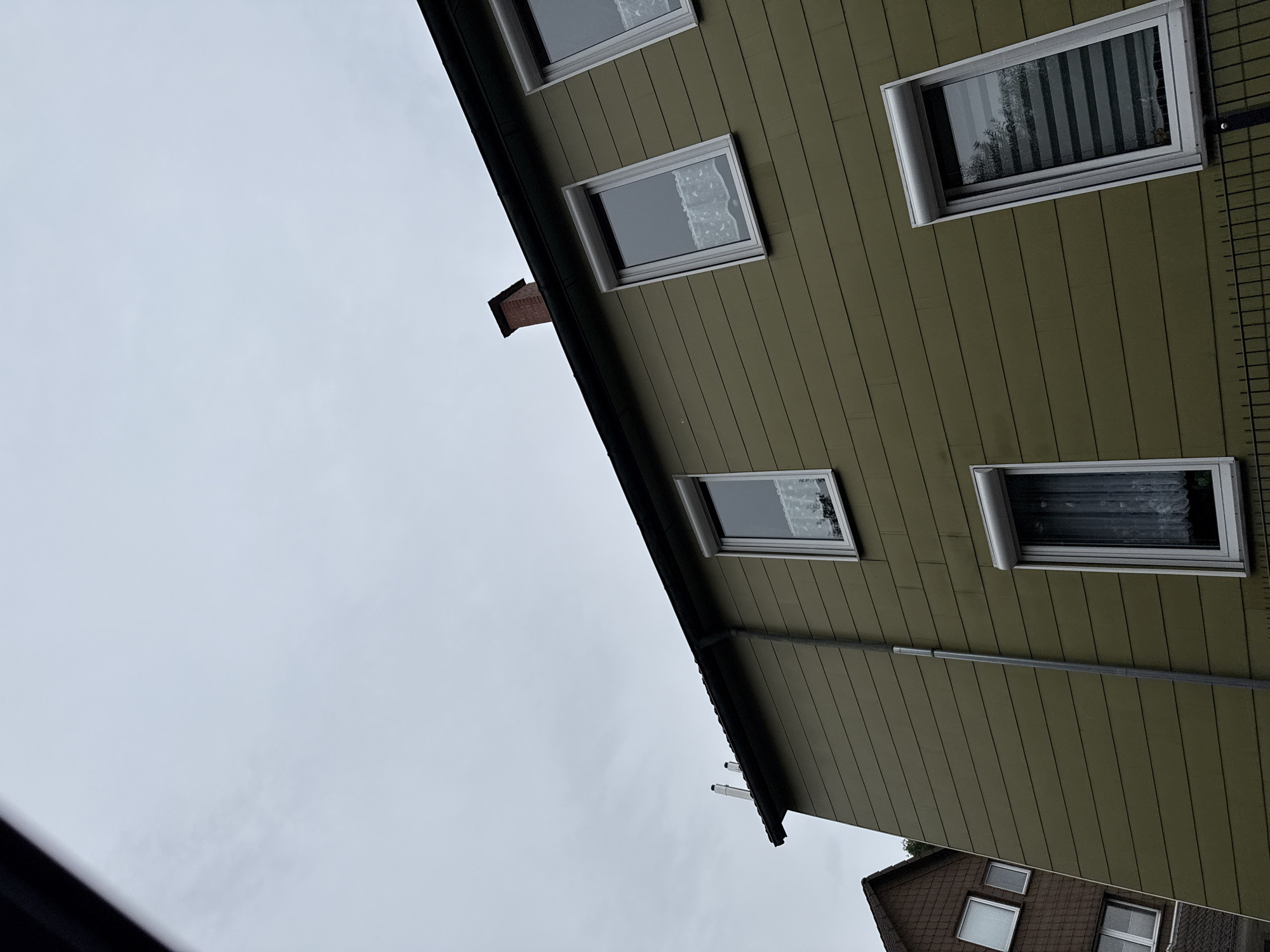 A low-angle, slightly tilted, full shot captures the exterior of a residential building in Bad Lauterberg im Harz, Germany, under an overcast sky. The building's facade is clad in horizontal olive-green siding. Several windows are visible, some with white roller shutters, and sheer white curtains are discernible behind the glass. One window displays a zebra blind. A brick chimney with four visible flues extends from the dark-colored roofline. To the left, the edge of another building with a brown, shingled facade and multiple windows is partially visible. A metal drainpipe runs vertically down the green siding. A black metal fence runs along the bottom of the frame. The overall impression is that of a residential street scene.