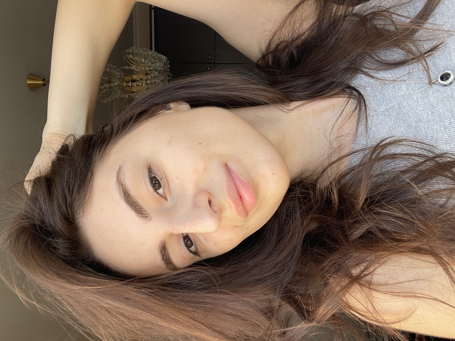 A close-up portrait depicts a single female subject with long, wavy, dark brown hair. Her right arm is raised, with her hand positioned above her head, appearing to run through or style her hair. She has fair skin, light brown eyes, and a subtle smile with slightly parted lips. Minor reddish marks are visible on her nose and cheeks. She is wearing a grey, ribbed tank top or camisole, accessorized with a silver-colored pendant featuring a large oval black stone, likely encircled by smaller clear stones, resting on her chest. The subject is looking directly at the camera. The background is softly out of focus, revealing elements of an indoor, residential setting. Visible objects include a gold-toned chandelier with crystal droplets in the upper right and dark brown furniture behind it. A brass-colored ceiling fixture is also partially discernible. The scene takes place within a domestic environment in the city of Moscow, Russia. No specific district or address is identifiable from the visual evidence.