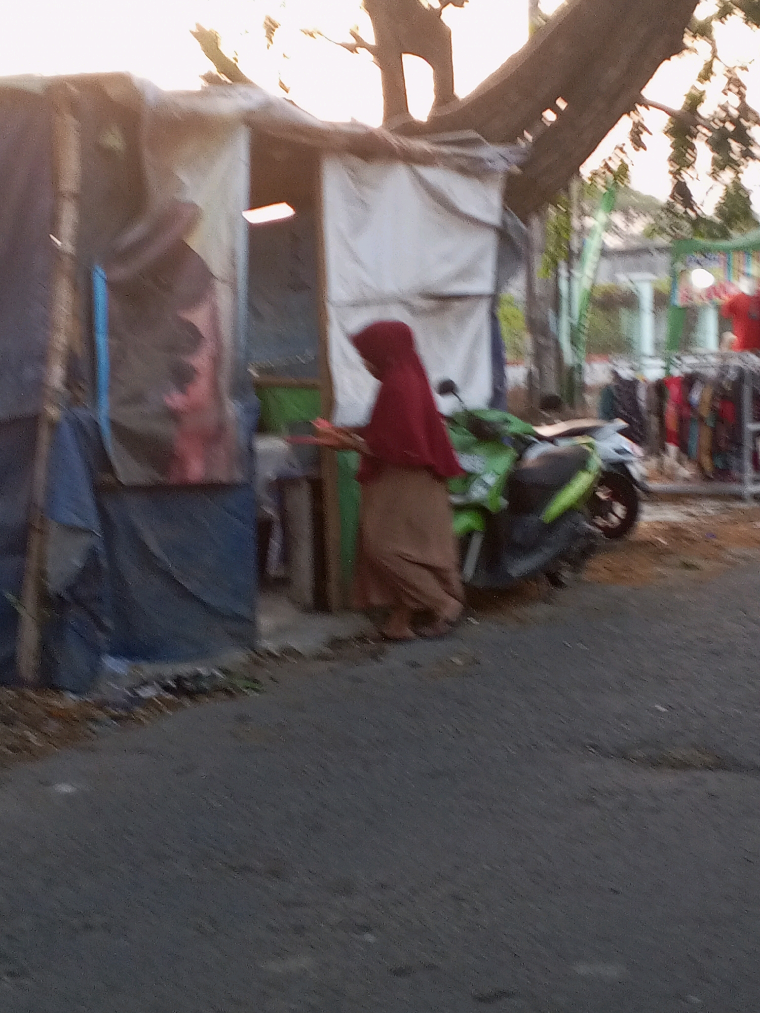 In Ranjeng, Indonesia, a person wearing a maroon hijab and a tan skirt stands next to a green motorcycle. The individual appears to be interacting with something on a small table or counter, which is partially obscured by the motorcycle. Behind them, a makeshift structure is visible, constructed from what appears to be tarps and fabric. A large tree branch extends over the scene from the upper right. In the background, more stalls with clothing displayed on racks suggest a marketplace or roadside vendor area. The ground in the foreground is asphalt, with some debris scattered along the edge. The lighting indicates it is either early morning or late afternoon.