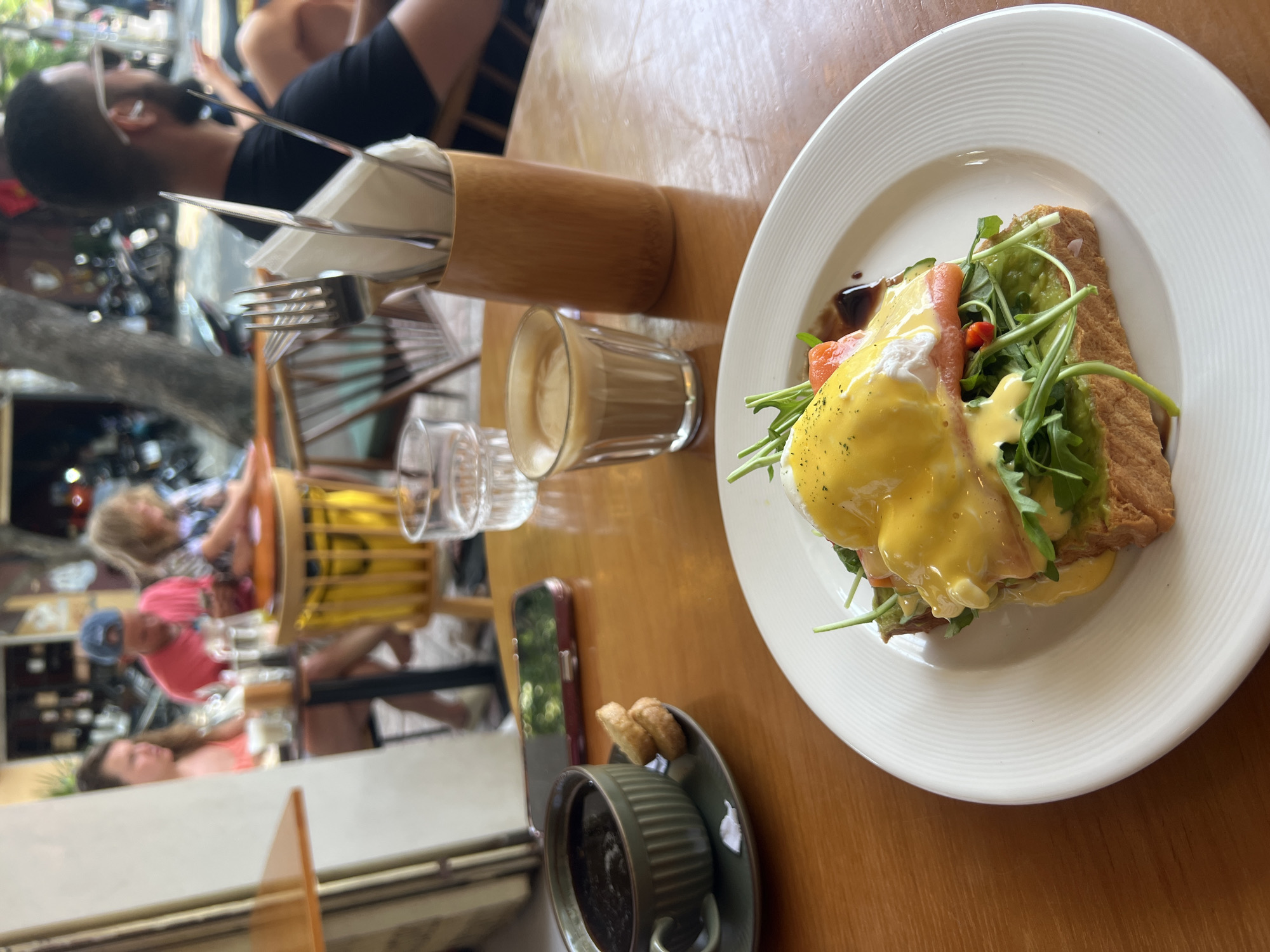 A high-angle indoor shot shows a table setting featuring a plate of Eggs Benedict with salmon and arugula on a wooden table, with other items including a bamboo cutlery holder with utensils, small glasses of milk or coffee, a cup of black coffee with cookies, and a smartphone. In the blurred background, visible through a window or open space, there are people, motorcycles, and a storefront, suggesting an outdoor scene in Nha Trang, Vietnam.