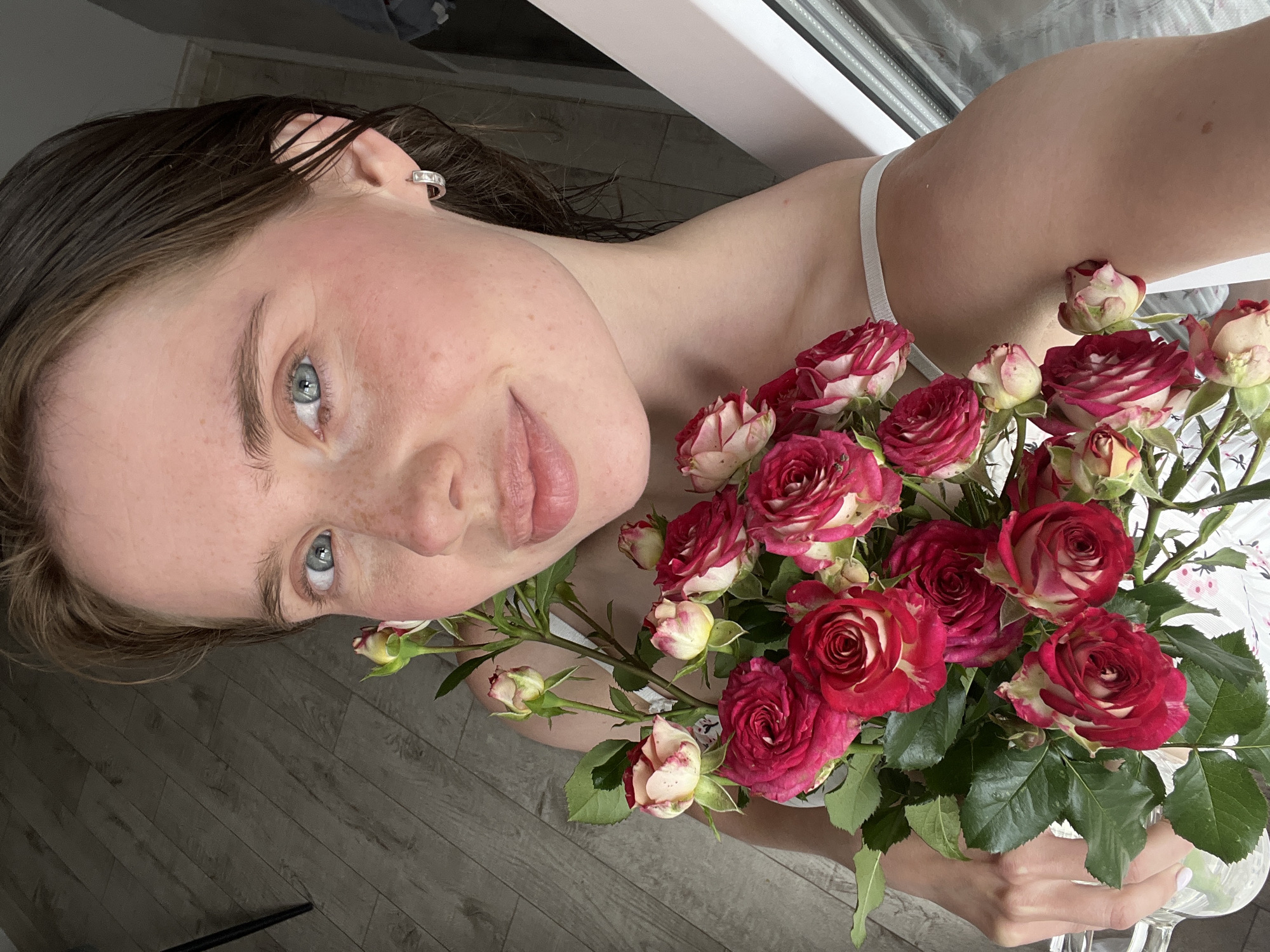 A young woman with light skin and light brown hair is holding a bouquet of roses. She is looking at the camera with a slight smile. Her eyes are blue and she has freckles across her nose and cheeks. She is wearing a white tank top and a silver hoop earring in her right ear. The roses are a mix of red and white. The background is a wooden floor and a white surface that appears to be a window frame or sill. The image appears to be taken indoors.