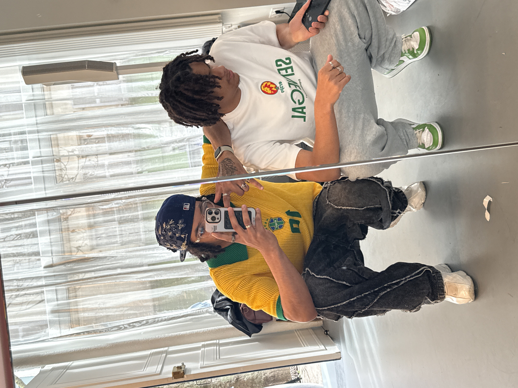 Two men are reflected in a mirror. The man in the foreground is wearing a yellow knit jersey with a green collar and a blue and floral cap. He is also wearing dark distressed pants and white sneakers. He is holding a phone in his hand, presumably taking the picture. The man in the background is wearing a white t-shirt and gray sweatpants. He is also wearing green and white sneakers. The scene is in an indoor setting, possibly a room or a studio with blinds on the window. There are no indicators of this being in Paris, France.