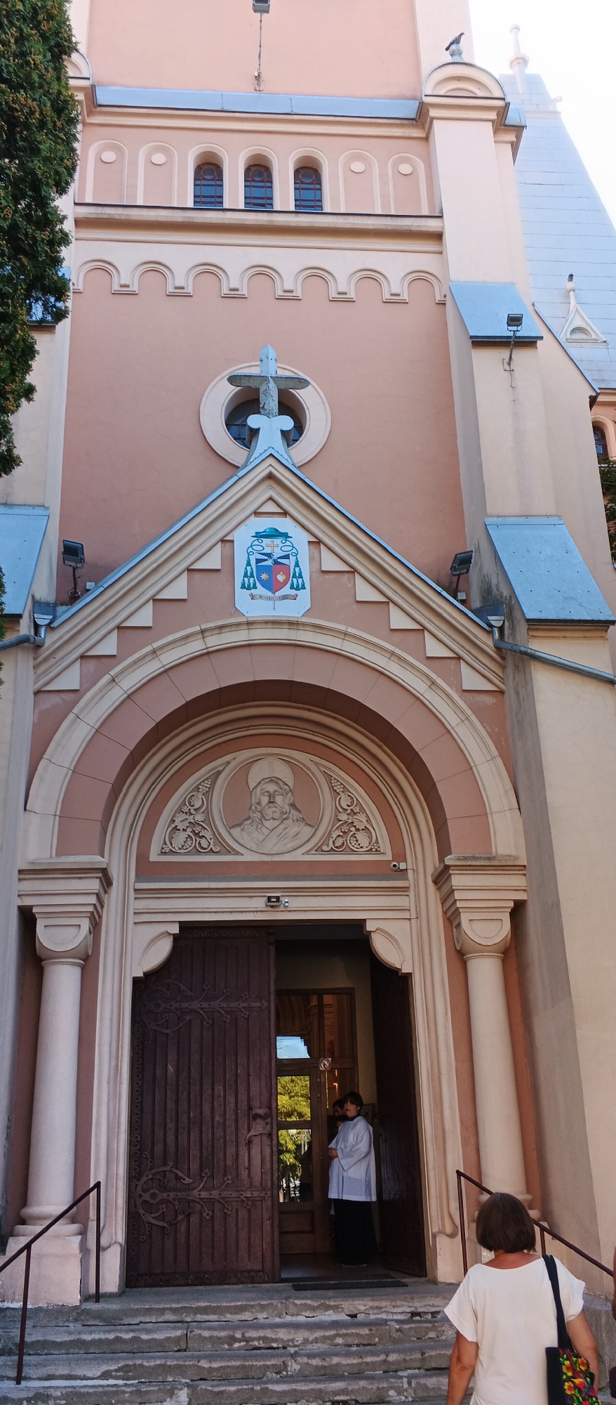 The image shows the exterior of the St. Martin's Cathedral in Mukachevo, Ukraine. The cathedral's facade is a pale pink color, with decorative arches and windows. Above the entrance, there is a large arched doorway with double wooden doors, one of which is open. Above the doorway is a relief carving of a head, likely depicting a saint or religious figure. A coat of arms is mounted on the gable above the relief. Two figures are visible at the entrance: one is wearing a white alb and a black skirt, standing just inside the doorway. Another person, a woman with short brown hair, is seen from behind walking up the stone steps towards the entrance, carrying a bag. The steps are made of rough stone and show signs of wear. To the left of the entrance, a large tree with dark green foliage is partially visible.