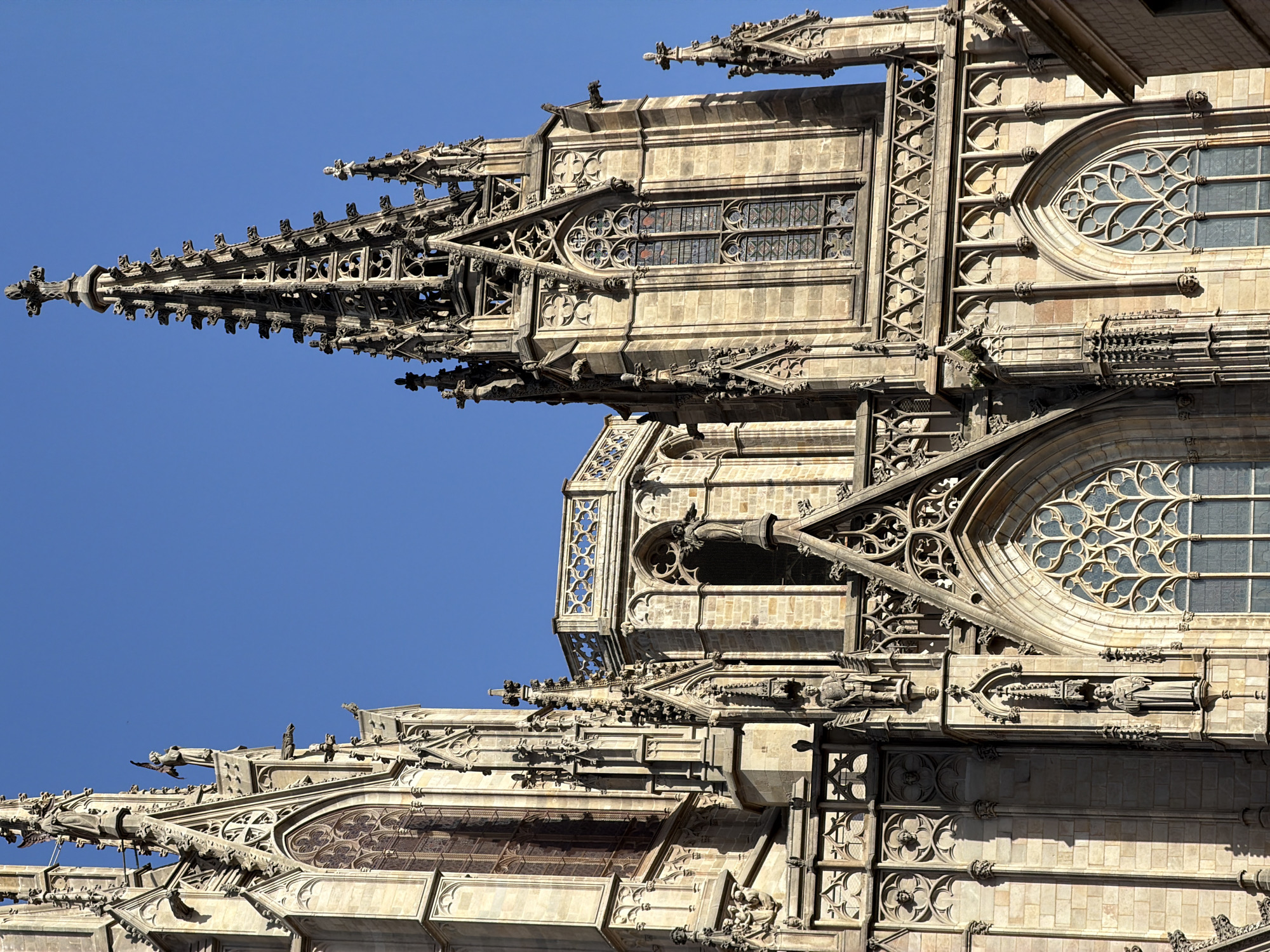 This image is a close-up, low-angle shot of the intricate Gothic architecture of a cathedral in Barcelona, Spain. The primary subjects are the stone structures, characterized by sharp spires, detailed tracery in the windows, and numerous gargoyles or decorative sculptures adorning the façade. The bright blue sky suggests a clear, sunny day, likely mid-morning or afternoon due to the strong sunlight casting shadows and highlighting the texture of the stone.
The cathedral features several levels of architectural complexity. The foreground shows the upper portions of the building with pointed arches, narrow stained-glass windows with complex patterns, and ornate stonework. In the mid-ground, a large, circular window with elaborate tracery is prominent. The spires and decorative elements extend outwards, creating a sense of depth and grandeur.
The stone is weathered, showing the age and history of the building. The gargoyles, positioned along ledges and corners, appear to be in various states of preservation, some more detailed than others. The overall impression is one of immense craftsmanship and architectural ambition. There is no visible text in the image. The scene evokes a sense of awe and wonder at the historical and artistic achievements of the cathedral's builders.