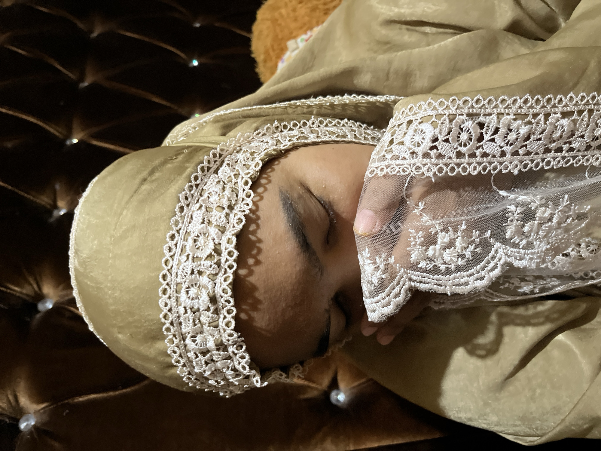 A close-up, vertical, medium shot shows a person wearing a golden hijab with intricate white lace trim around the forehead and veil. The person's eyes are closed, and their hands, also adorned with white lace, are held up to their face, covering their mouth and nose. The person is seated on a dark brown, tufted couch, with hints of a fuzzy orange object visible behind them. The lighting is dim, creating shadows and highlighting the texture of the fabric and lace. There is no visible indication of the location being in Kruengmate, Indonesia, or any other specific city.